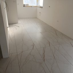 LM Wykończenia Wnętrz - Jasne, marmurowe płytki podłogowe z brązowymi żyłkami w nowym wnętrzu. Widoczne kable elektryczne i przygotowane otwory na gniazdka. Okno zabezpieczone folią.