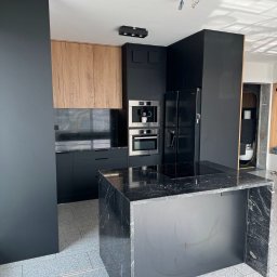 LM Wykończenia Wnętrz - Nowoczesna kuchnia z wyspą wykończoną czarnym marmurem, drewniane szafki górne, czarne szafki dolne i sprzęt AGD. Na podłodze szare płytki, w rogu widoczna skrzynka z narzędziami.