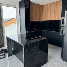 LM Wykończenia Wnętrz - Nowoczesna kuchnia z wyspą z czarnego granitu, drewnianymi szafkami i czarnymi frontami. Widoczne okno i narzędzia, sugerujące etap wykończenia.