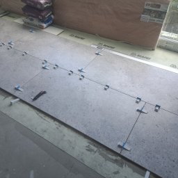 LM Wykończenia Wnętrz - Układanie szarych płytek z użyciem systemu poziomowania. Widoczne worki z klejem, poziomica i nóż. Prace glazurnicze w trakcie realizacji, z bliska.