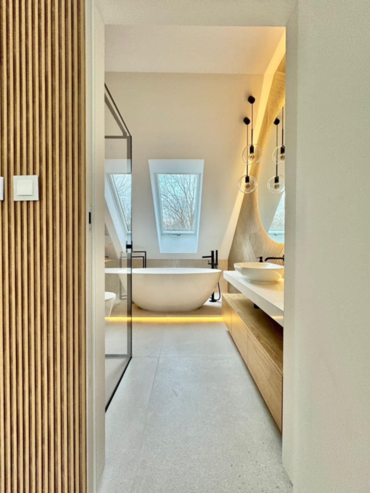 Nowoczesna łazienka na poddaszu z wolnostojącą wanną, oświetleniem LED i drewnianymi lamelami przy wejściu. Minimalistyczny design z oknem dachowym.