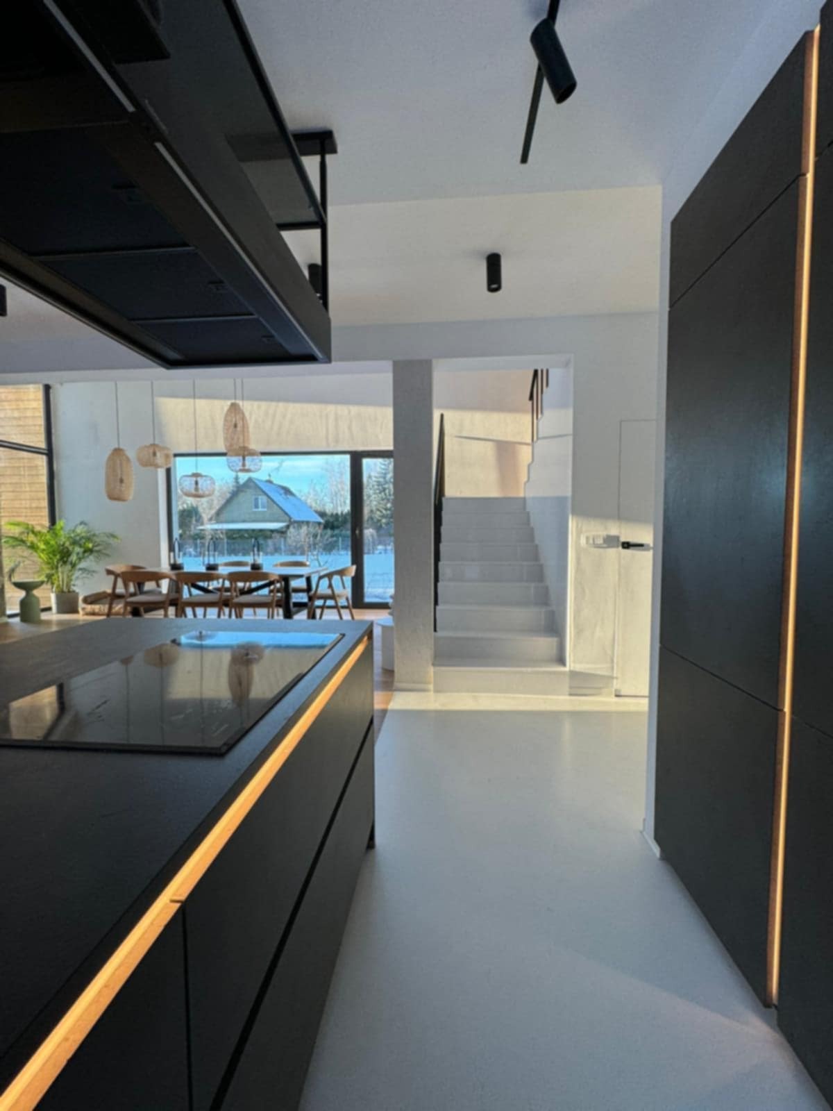 Nowoczesna kuchnia z wyspą i oświetleniem LED, widok na jadalnię i schody. Minimalistyczny design, jasne wnętrze z dużymi oknami i widokiem na zimowy krajobraz.