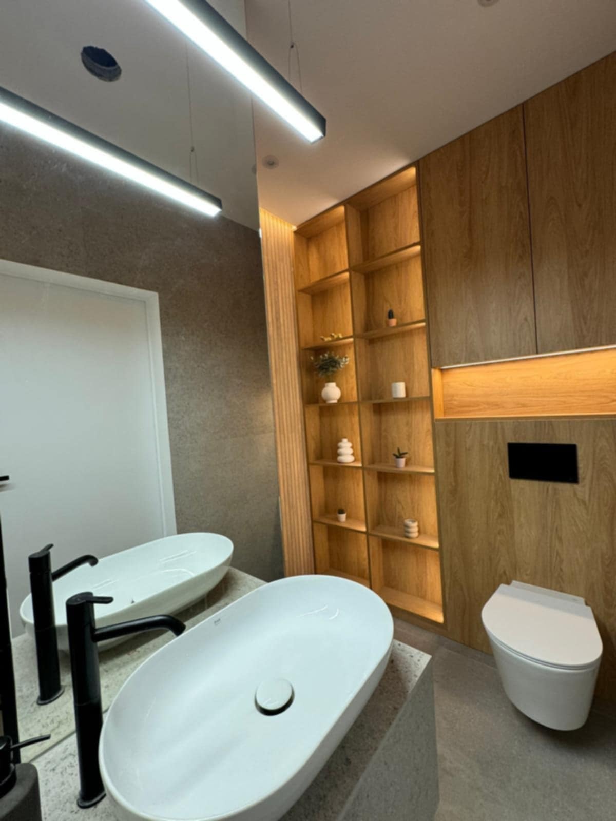 Nowoczesna łazienka z dwiema owalnymi umywalkami na kamiennym blacie, drewnianą zabudową z półkami i podświetleniem oraz wiszącą toaletą.