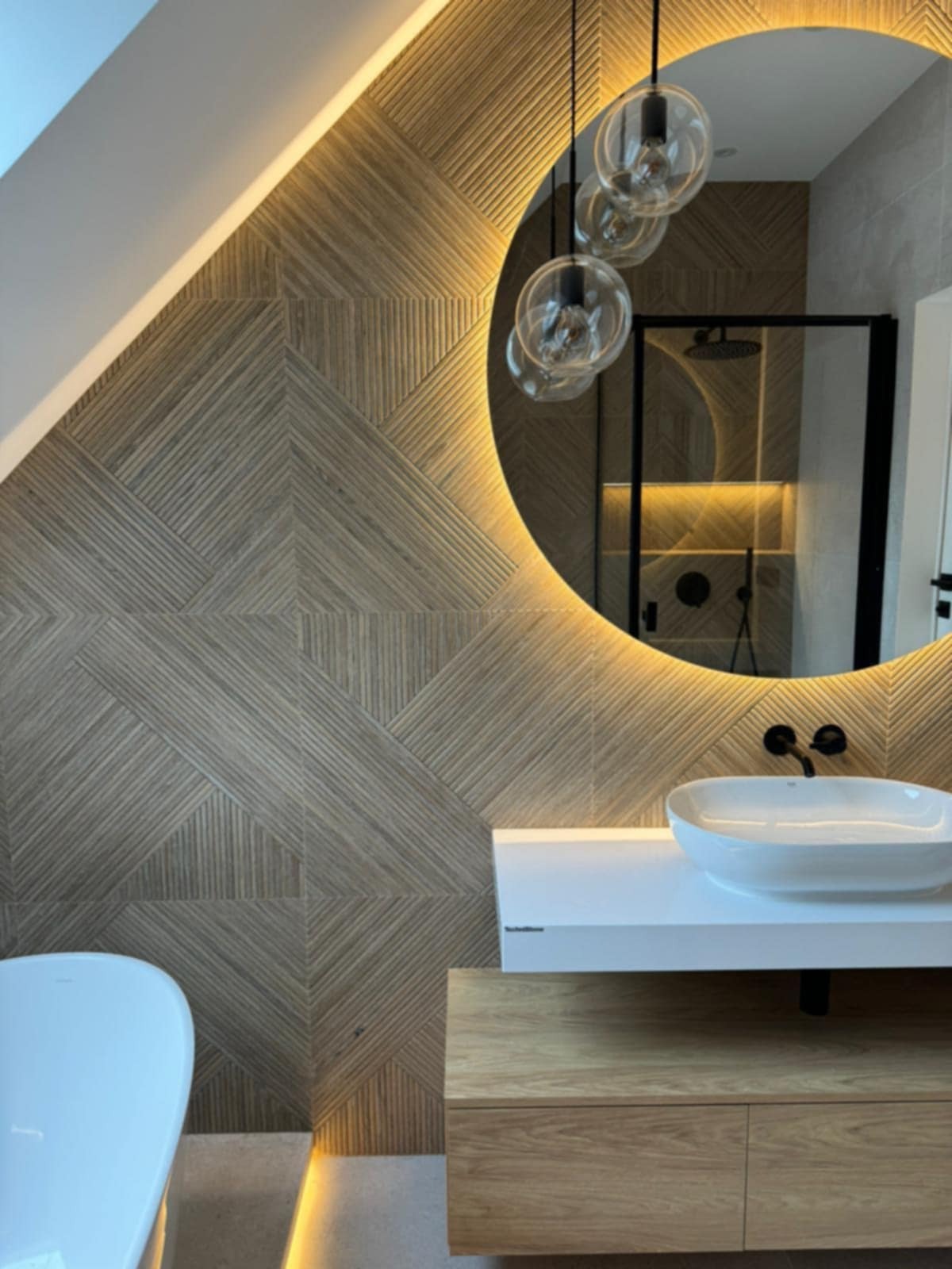 Nowoczesna łazienka z drewnianą ścianą w geometryczne wzory, okrągłym lustrem z podświetleniem i wiszącymi lampami. Widoczna umywalka i fragment wanny.