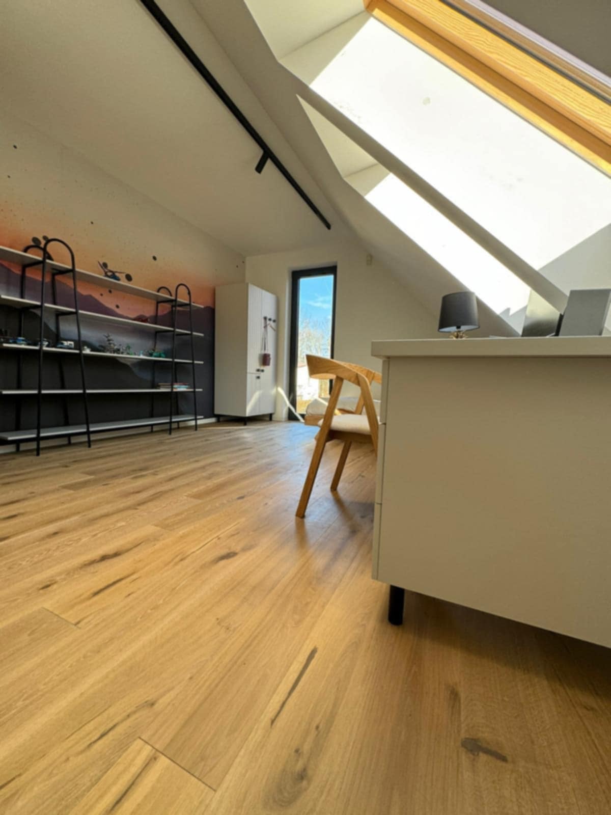 Jasna drewniana podłoga w pokoju na poddaszu z oknem dachowym, biurkiem i regałem na ścianie. Widoczne naturalne usłojenie drewna i minimalistyczny design wnętrza.