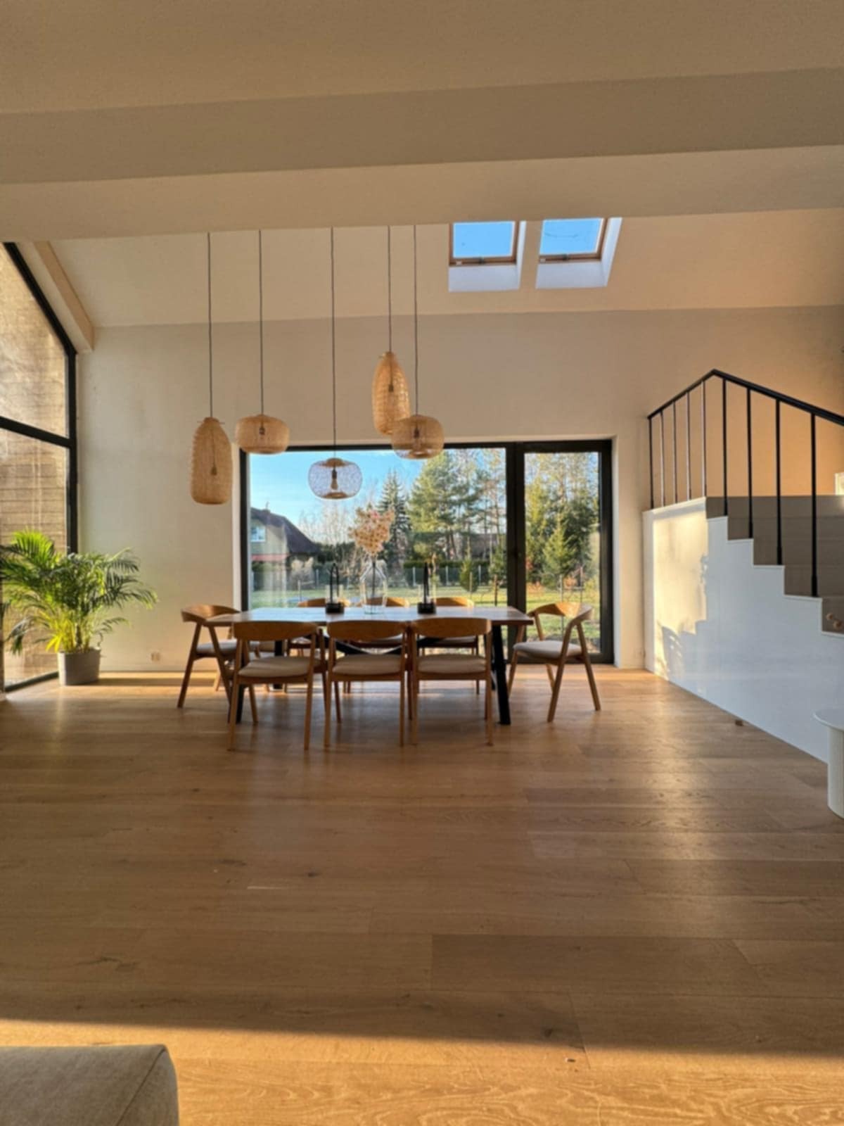 Jasne, przestronne wnętrze z drewnianą podłogą, stołem jadalnym i schodami. Naturalne światło wpada przez okna dachowe i duże okno z widokiem na zieleń.