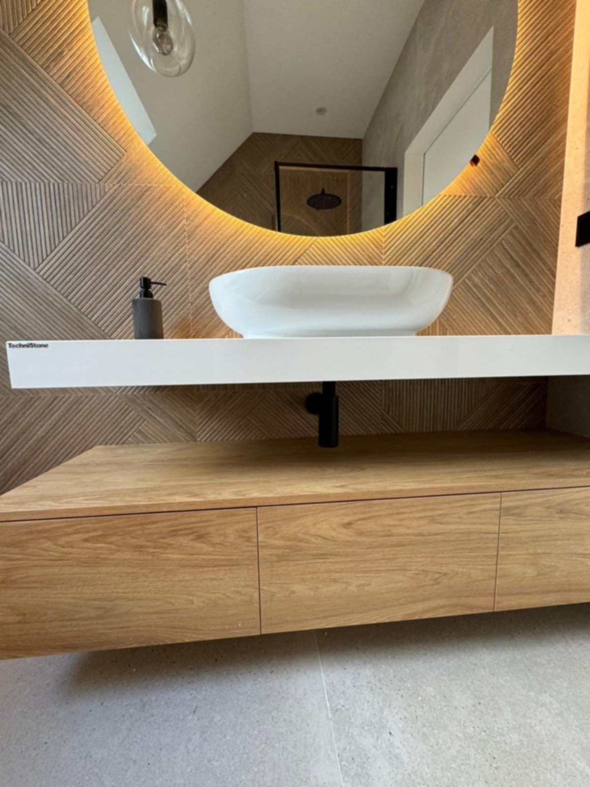 Nowoczesna łazienka z okrągłym lustrem podświetlanym LED, umywalką nablatową i drewnianą szafką. Dekoracyjne płytki na ścianie z geometrycznym wzorem.