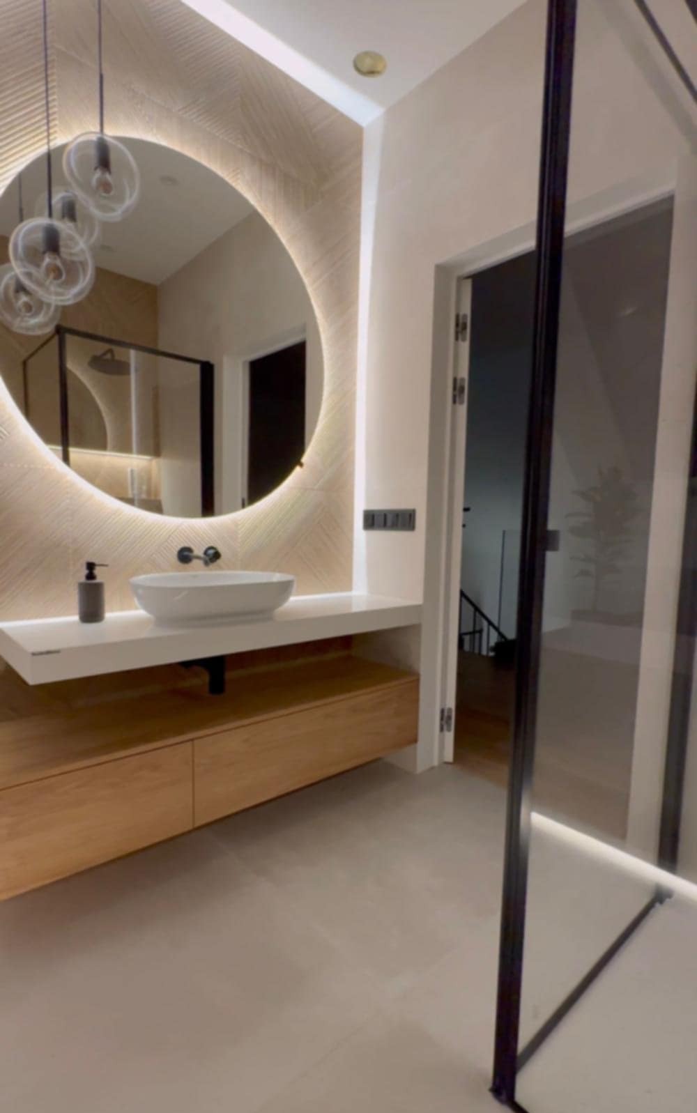 Nowoczesna łazienka z okrągłym lustrem podświetlanym LED, białą umywalką nablatową i drewnianą szafką pod umywalką. Widoczna kabina prysznicowa i wejście do pomieszczenia.