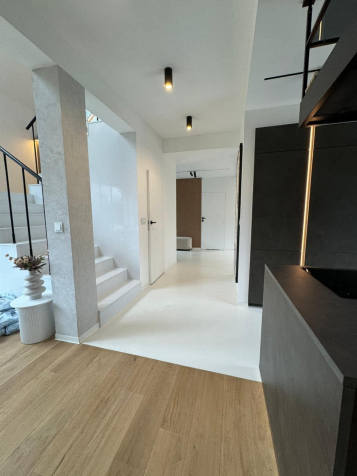 Nowoczesne wnętrze domu z drewnianą podłogą, białymi schodami i minimalistyczną kuchnią z czarnymi szafkami. Widoczne oświetlenie punktowe i dekoracyjny wazon.