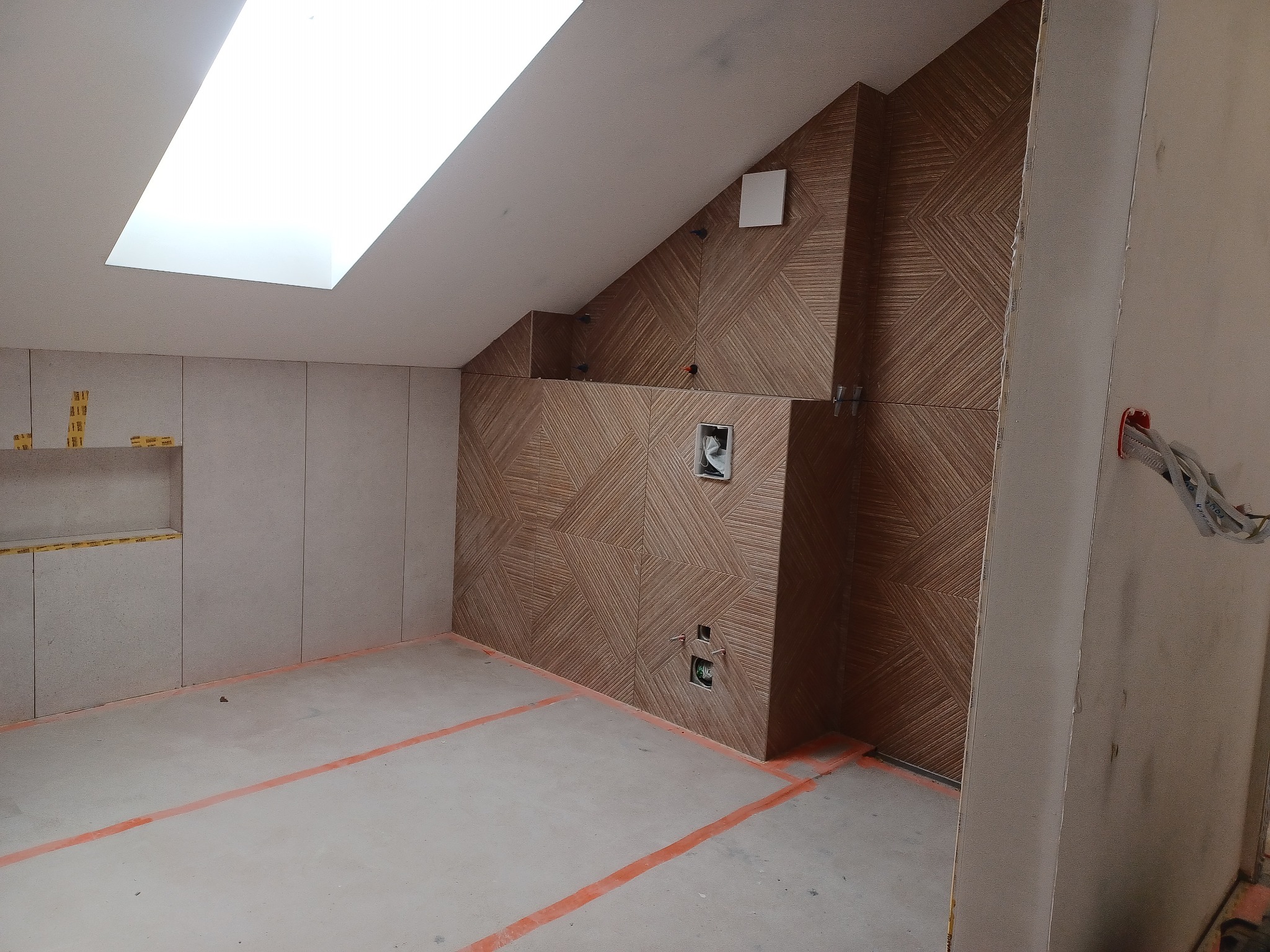 Wykończona łazienka na poddaszu z brązowymi płytkami ułożonymi w geometryczny wzór na ścianie. Widoczne przygotowane punkty instalacji i okno dachowe.