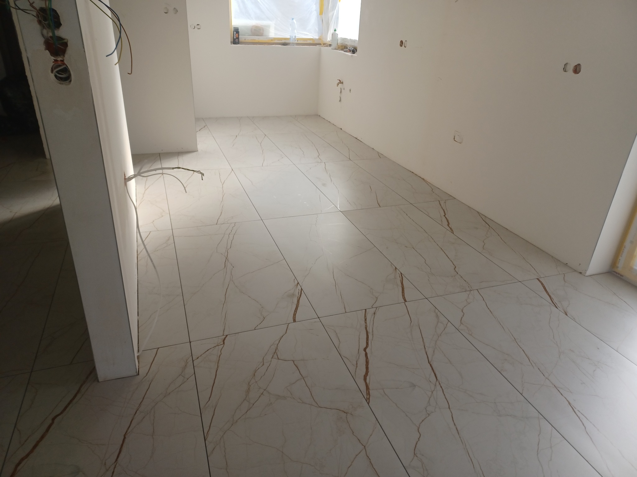 Jasne, marmurowe płytki podłogowe z brązowymi żyłkami w nowym wnętrzu. Widoczne kable elektryczne i przygotowane otwory na gniazdka. Okno zabezpieczone folią.