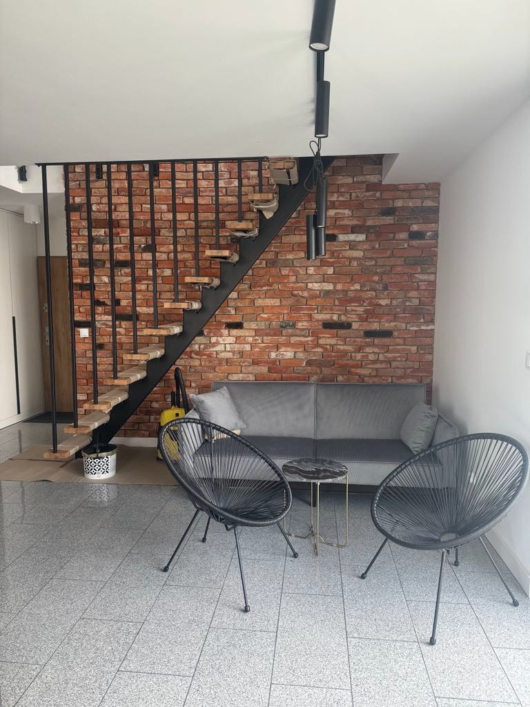 Salon z ceglaną ścianą, schodami z czarną balustradą, szarą sofą i dwoma czarnymi krzesłami. Na podłodze szare płytki. Widoczny odkurzacz.