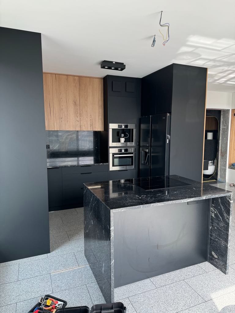 Nowoczesna kuchnia z wyspą wykończoną czarnym marmurem, drewniane szafki górne, czarne szafki dolne i sprzęt AGD. Na podłodze szare płytki, w rogu widoczna skrzynka z narzędziami.