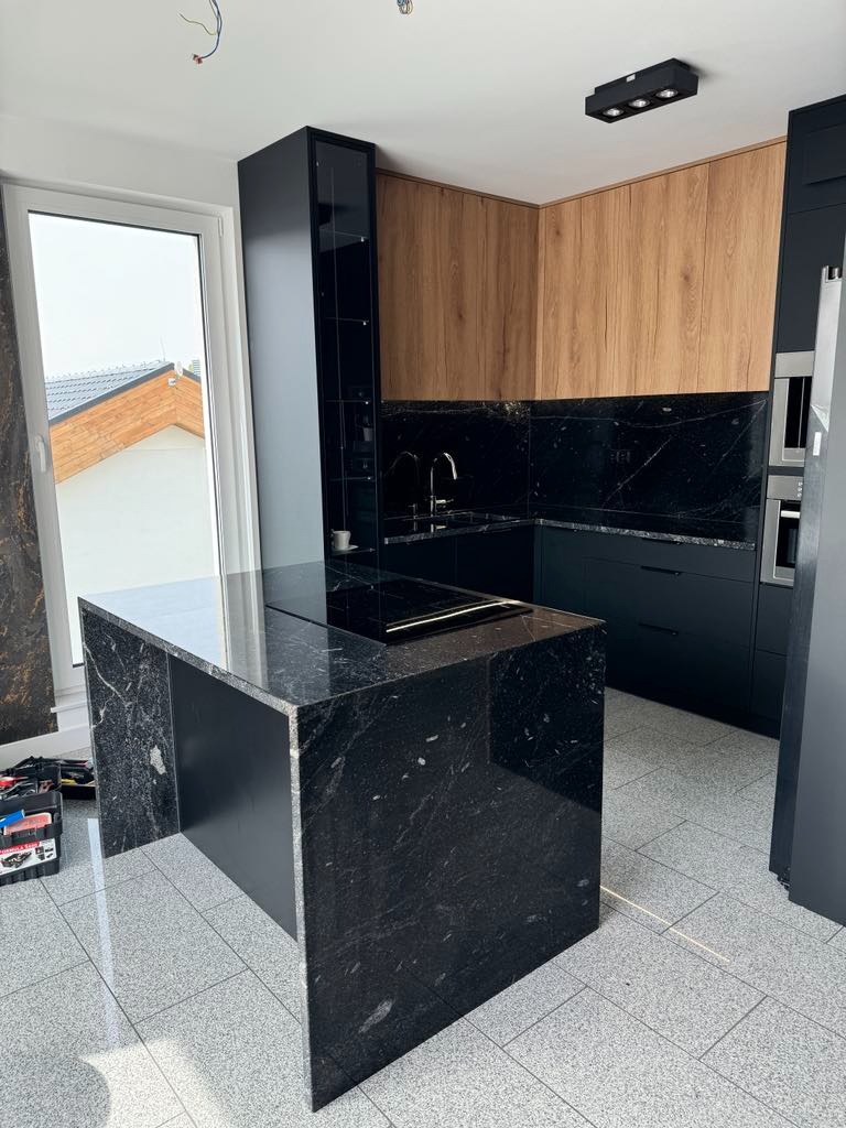 Nowoczesna kuchnia z wyspą z czarnego granitu, drewnianymi szafkami i czarnymi frontami. Widoczne okno i narzędzia, sugerujące etap wykończenia.