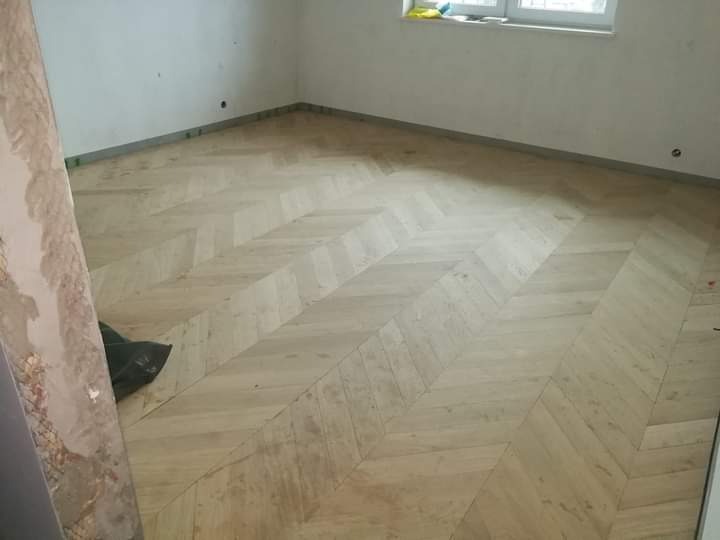 Jasny parkiet ułożony we wzór jodełki w pustym pokoju, widok z góry. Fragment ściany z resztkami kleju po lewej stronie. Okno w tle.