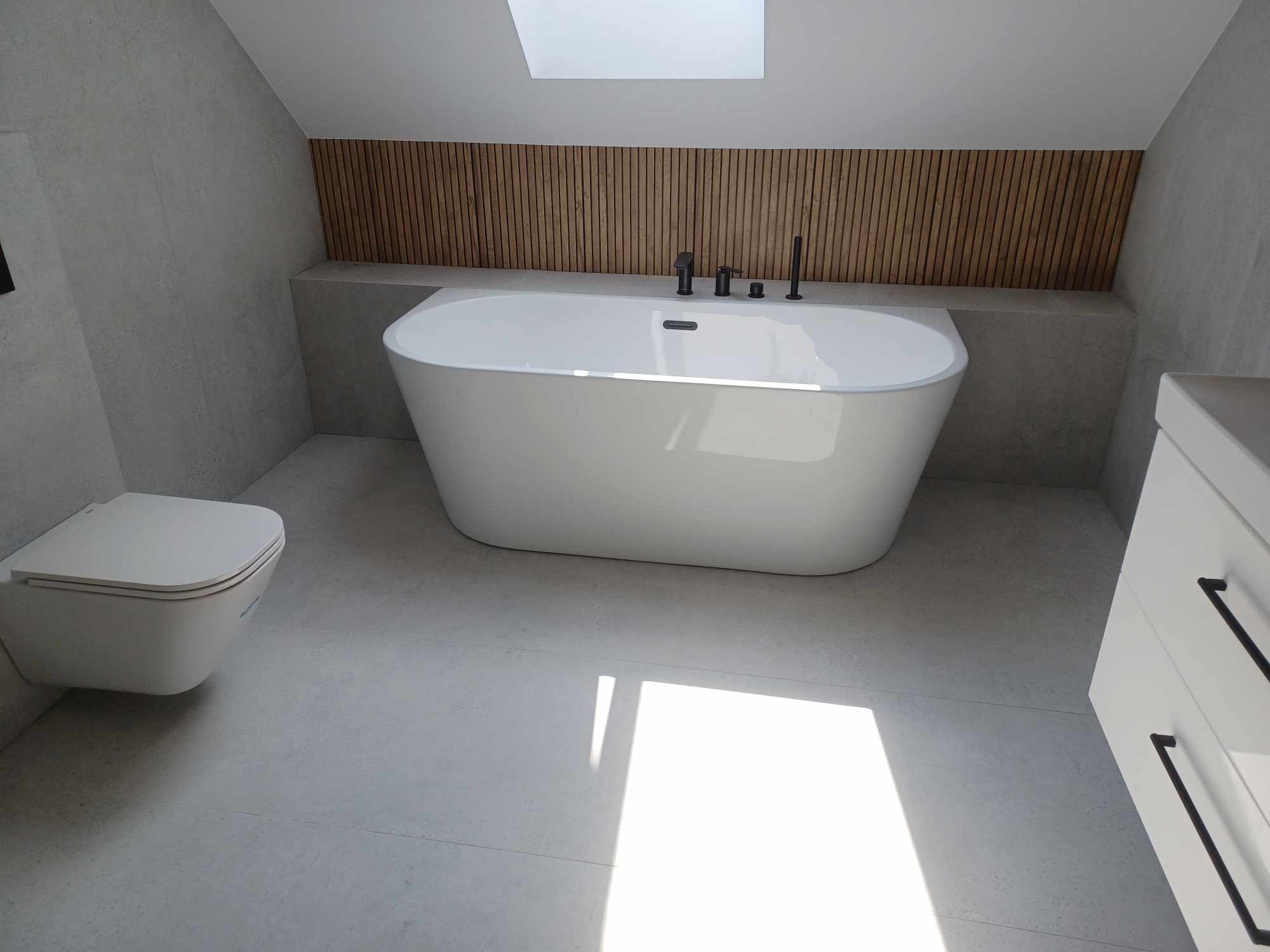 Nowoczesna łazienka na poddaszu z wolnostojącą wanną, drewnianym panelem dekoracyjnym i minimalistyczną toaletą. Jasne płytki i wpadające światło podkreślają przestronność.