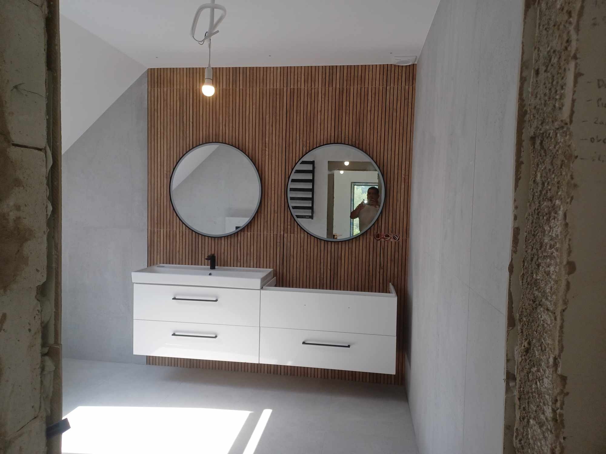 Nowoczesna łazienka w trakcie wykończenia: biała szafka pod umywalką, okrągłe lustra na tle drewnianej ściany, surowy beton w tle. Widoczne ślady prac remontowych.