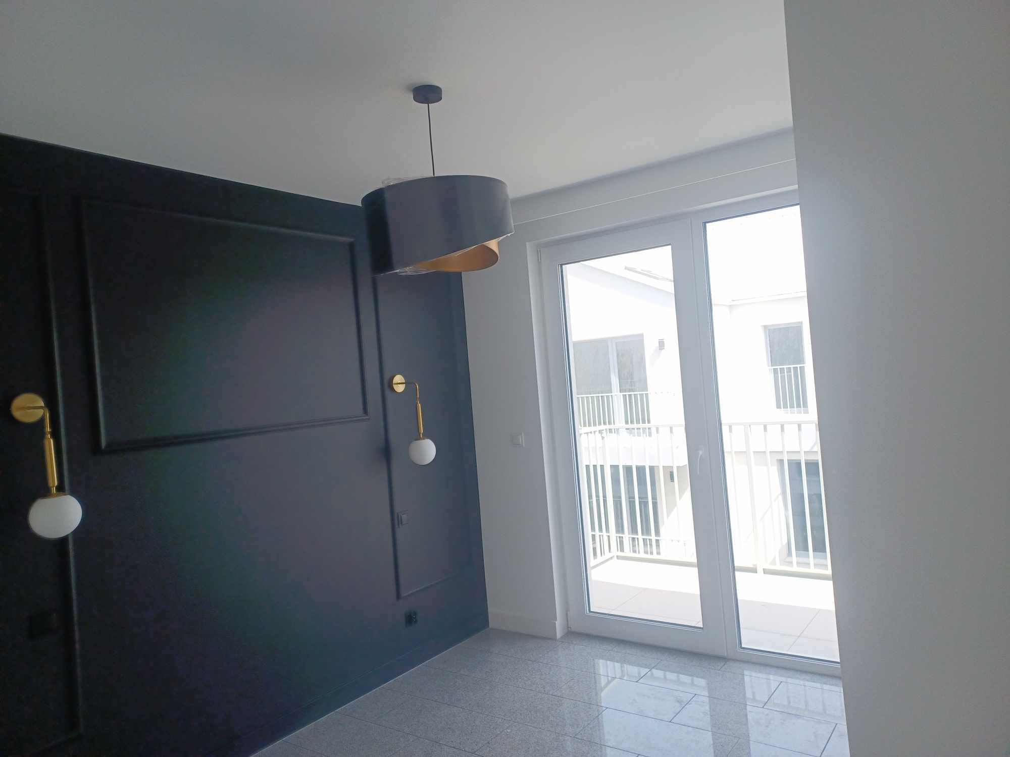 Eleganckie wnętrze: ciemna ściana z dekoracyjnymi listwami, złote kinkiety z kulistymi kloszami, nowoczesna lampa wisząca i jasne okno balkonowe.