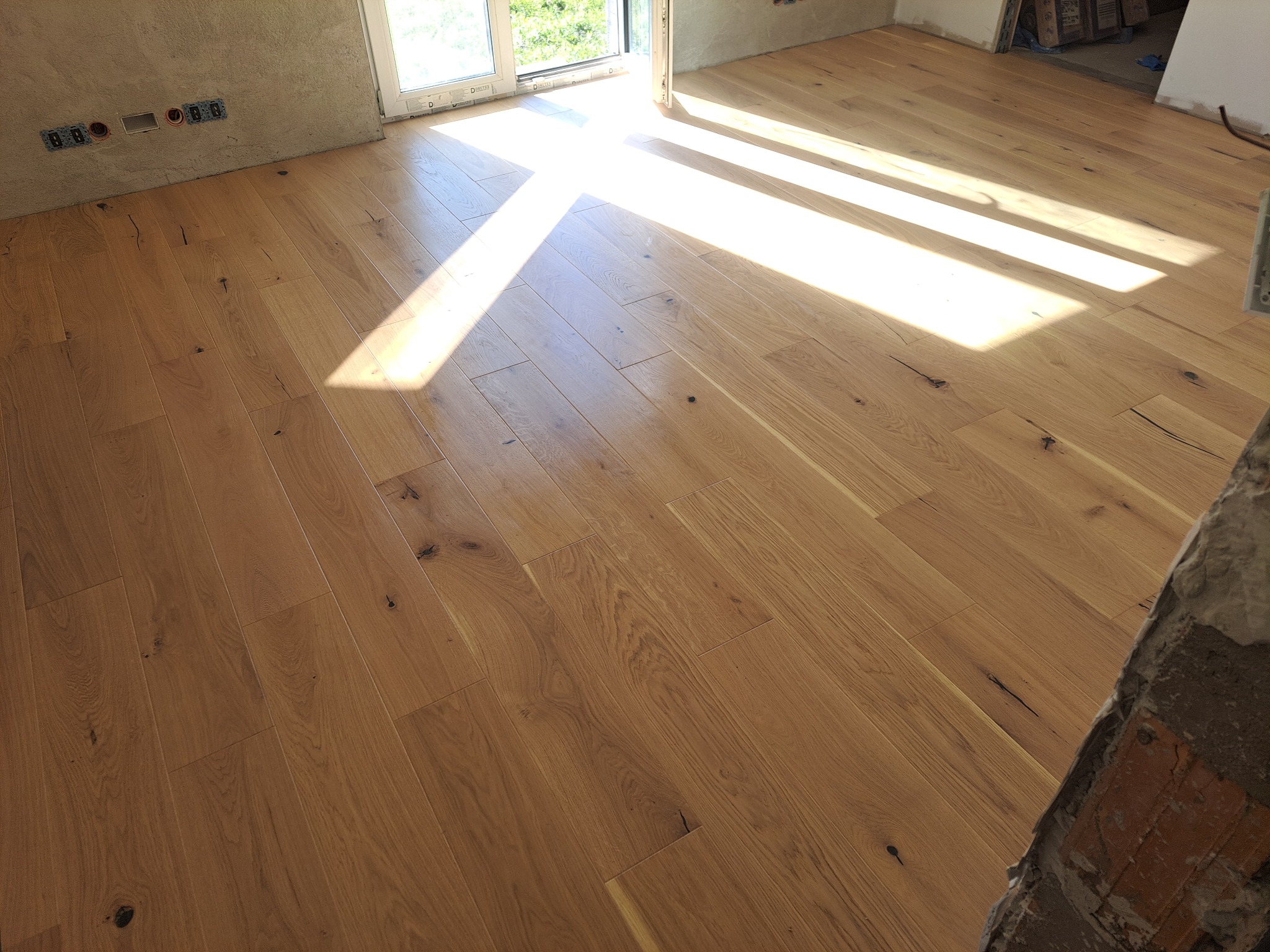Jasna, dębowa podłoga ułożona w jodełkę w Głoskowie, z widocznymi promieniami słońca wpadającymi przez okno. Ściany przygotowane do dalszych prac wykończeniowych.