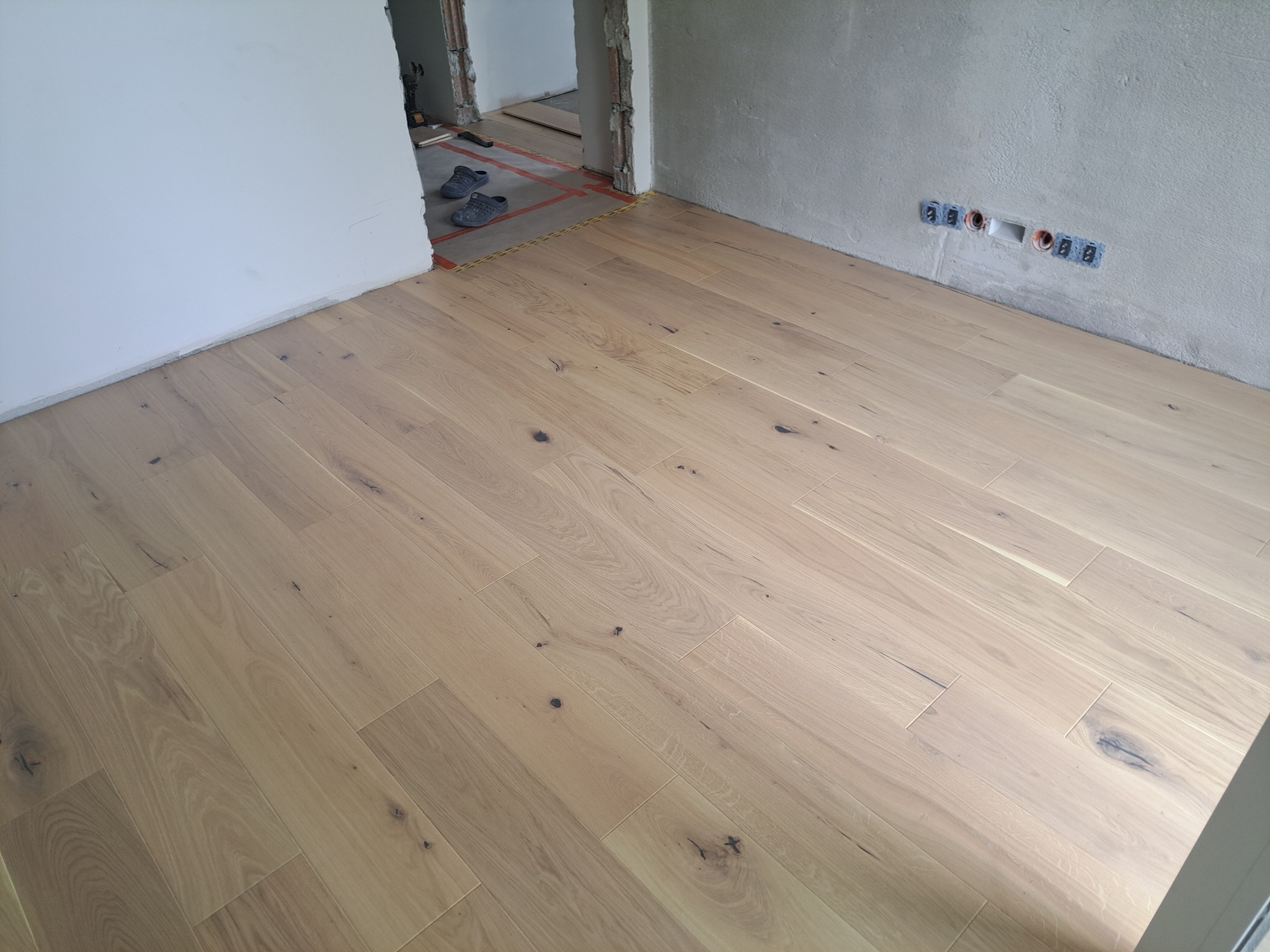 Jasny parkiet ułożony w pokoju z surowymi ścianami i widocznymi gniazdkami elektrycznymi. W tle widoczne buty robocze i narzędzia, co sugeruje trwający remont.