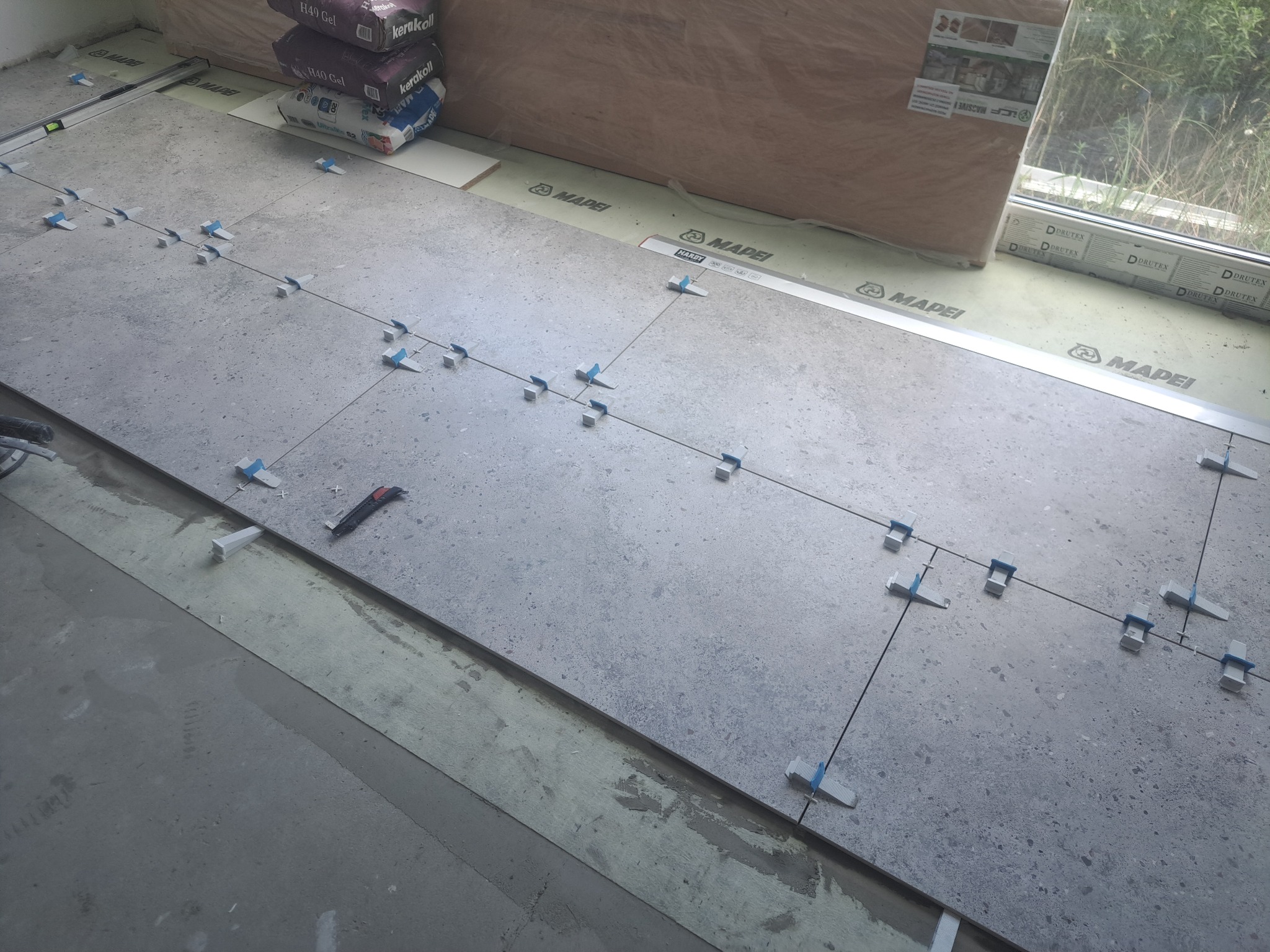 Układanie szarych płytek z użyciem systemu poziomowania. Widoczne worki z klejem, poziomica i nóż. Prace glazurnicze w trakcie realizacji, z bliska.