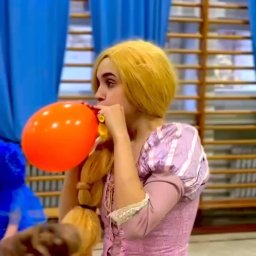 IG Artist Agency - Animator w stroju Roszpunki dmucha pomarańczowy balon na tle sali gimnastycznej z niebieskimi zasłonami. Widoczny fragment kostiumu i warkocz.