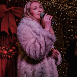 IG Artist Agency - Blond piosenkarka w futrze śpiewa do mikrofonu na tle czerwonej dekoracji i światełek, tworząc świąteczny klimat występu muzycznego.