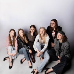 IG Artist Agency - Siedząca grupa sześciu młodych kobiet w różnorodnych stylizacjach, uśmiechniętych, na jasnoszarym tle. Zdjęcie studyjne, dobrze oświetlone, z widocznymi detalami ubrań i obuwia.