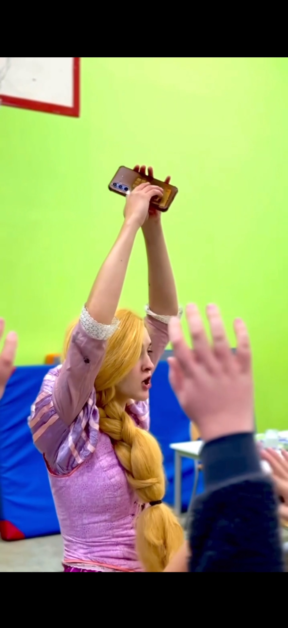 Animator w stroju Roszpunki trzyma telefon nad głowami dzieci podczas animacji. Jasnozielone ściany, sala zabaw.