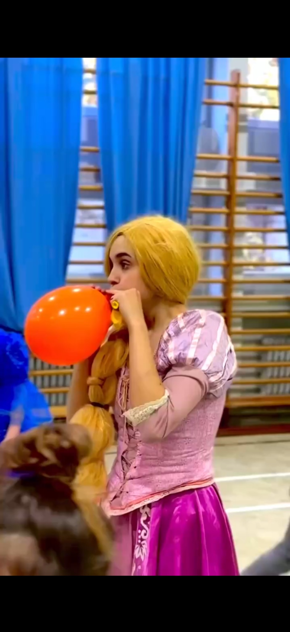 Animator w stroju Roszpunki dmucha pomarańczowy balon na tle sali gimnastycznej z niebieskimi zasłonami. Widoczny fragment kostiumu i warkocz.