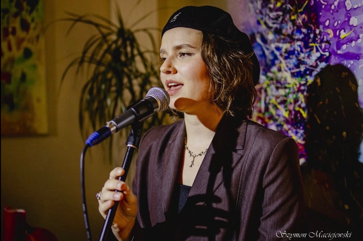 Wokalistka w brązowym garniturze i berecie śpiewa do mikrofonu na tle abstrakcyjnego obrazu. Scena oświetlona ciepłym światłem.