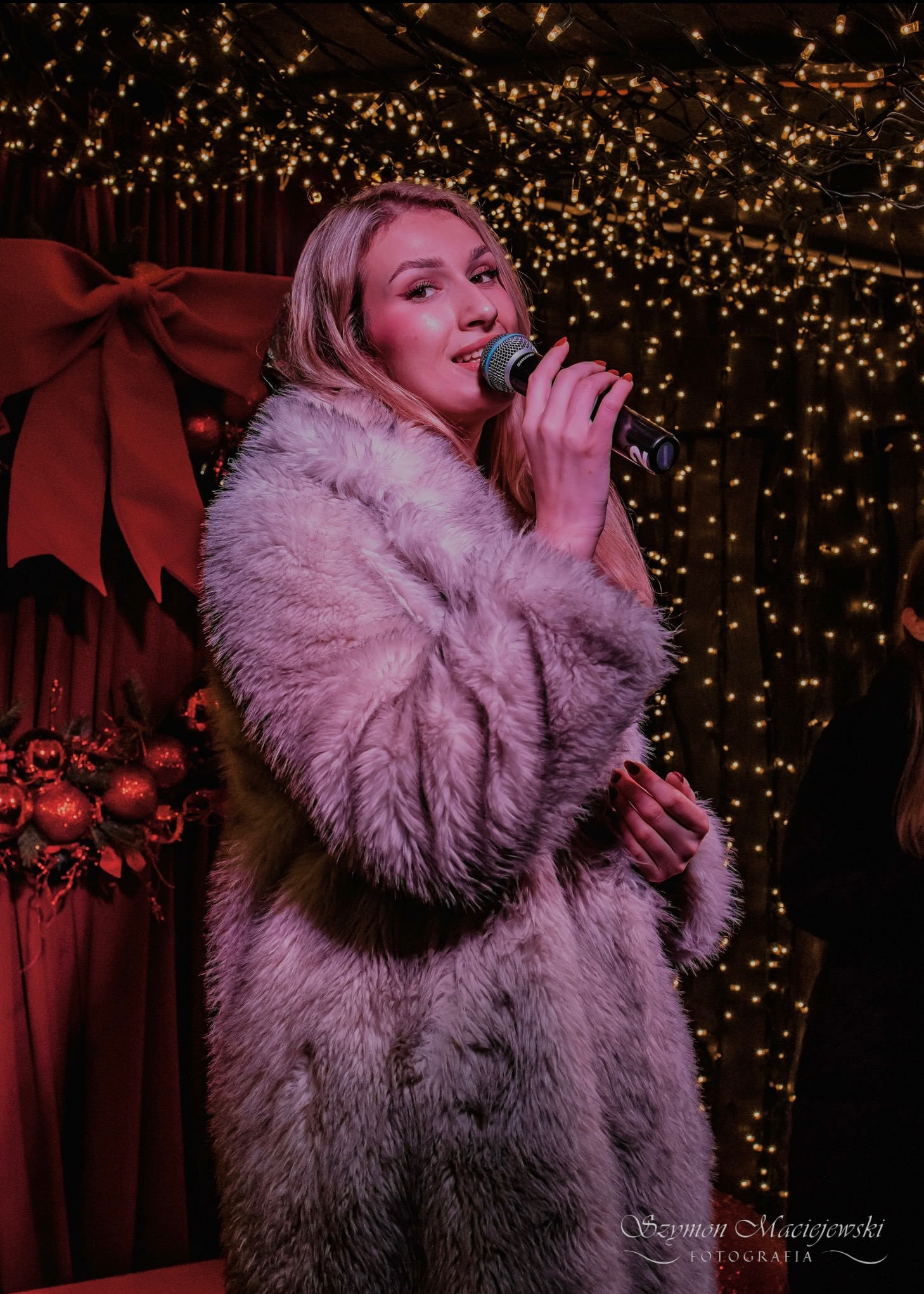 Blond piosenkarka w futrze śpiewa do mikrofonu na tle czerwonej dekoracji i światełek, tworząc świąteczny klimat występu muzycznego.