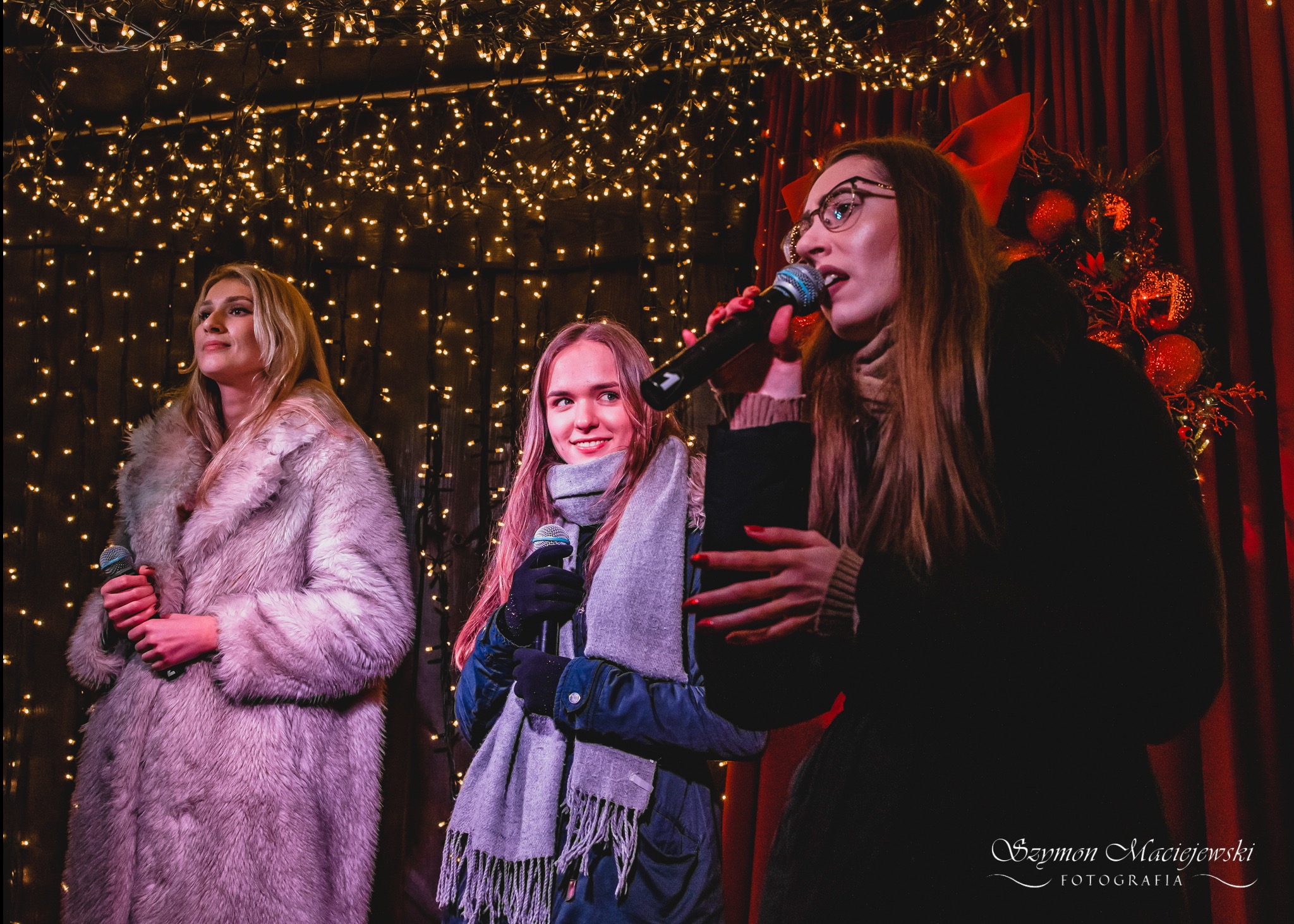 Trzy wokalistki z mikrofonami śpiewają na scenie z dekoracją świetlną i czerwoną zasłoną w tle. Jedna z nich ma okulary.
