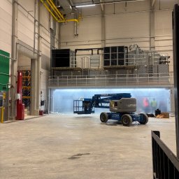 ICOOTECH SPÓŁKA Z OGRANICZONĄ ODPOWIEDZIALNOŚCIĄ - Montaż przemysłowej klimatyzacji w hali: podnośnik koszowy przed dużym agregatem, widoczne żółte rury wentylacyjne pod sufitem. Hala w trakcie prac instalacyjnych.