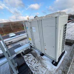 ICOOTECH SPÓŁKA Z OGRANICZONĄ ODPOWIEDZIALNOŚCIĄ - Biała, zewnętrzna jednostka klimatyzacji Daikin na dachu budynku, widoczna z perspektywy drabiny. Na dachu resztki śniegu, w tle budynek przemysłowy i błękitne niebo.