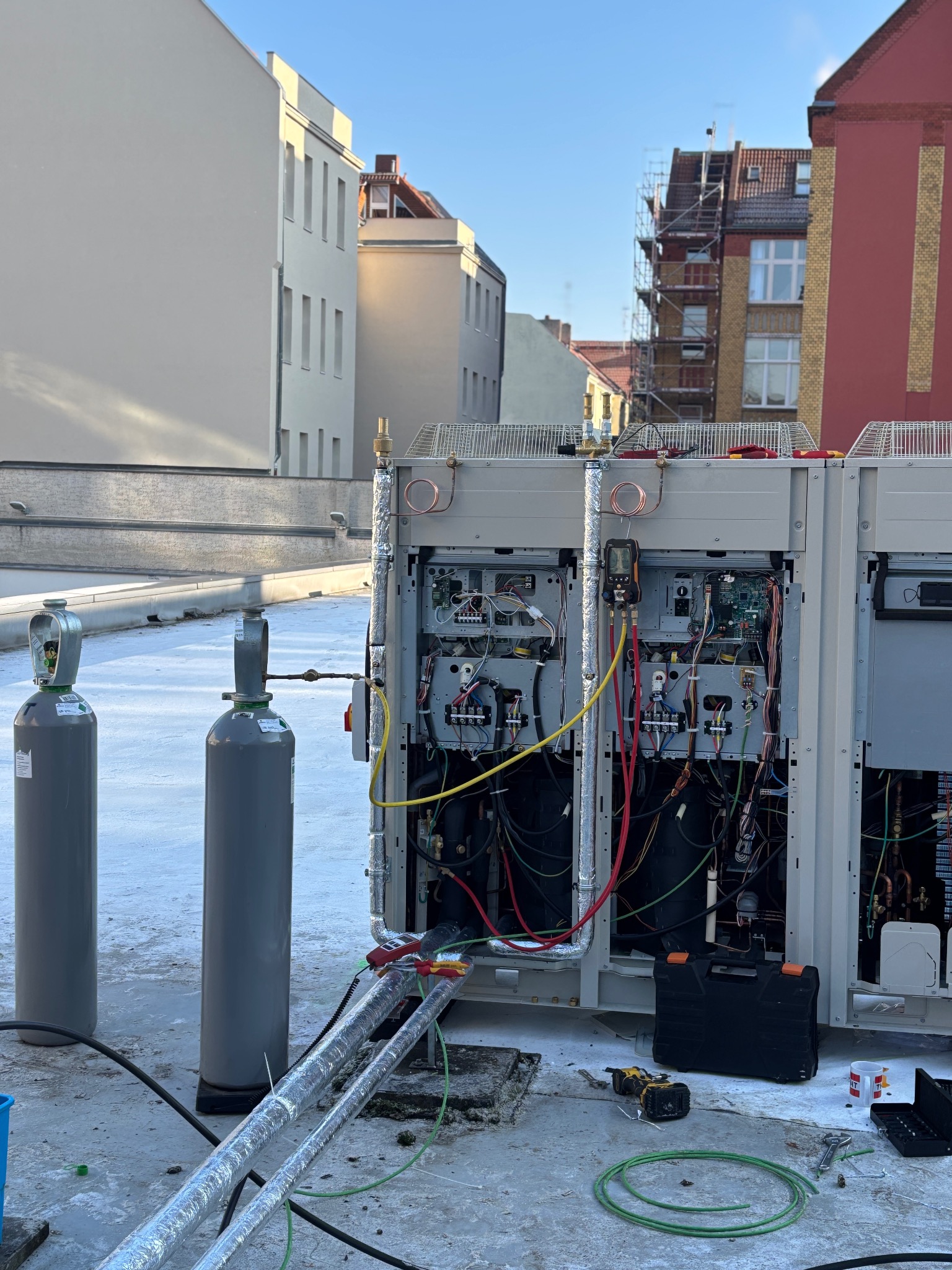 Instalacja klimatyzacji na dachu budynku, widoczne butle z gazem, narzędzia i otwarte panele urządzenia z okablowaniem. W tle zabudowa miejska i błękitne niebo.