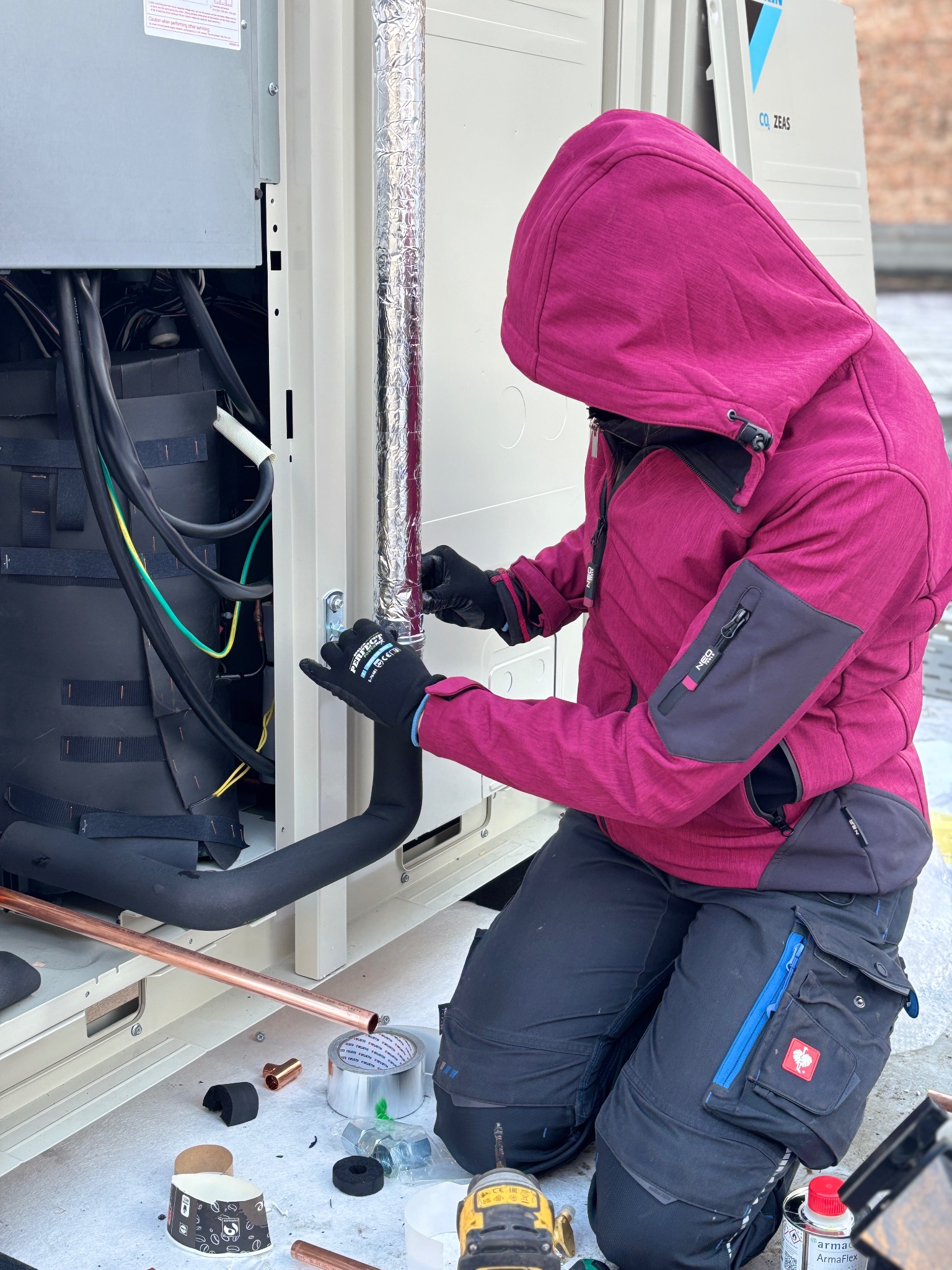 Instalator w różowej kurtce montuje rurę klimatyzacji. Widoczne narzędzia, miedziane rury i izolacja. Praca na zewnątrz, jasne światło dzienne.