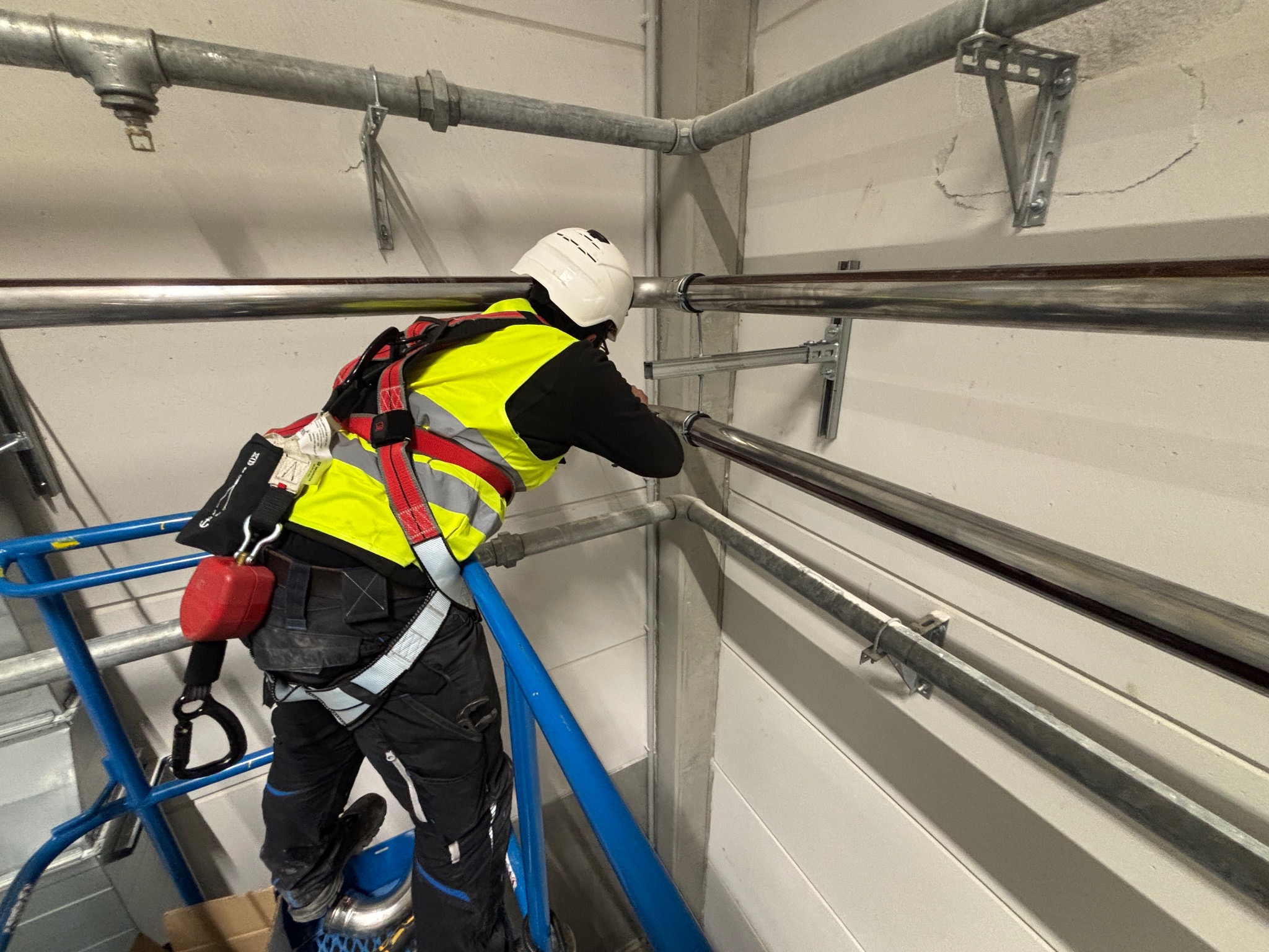 Montaż rur instalacyjnych pod sufitem przez pracownika w Gąsawie. Widoczny podnośnik i elementy mocujące rury do ściany. Praca na wysokości.