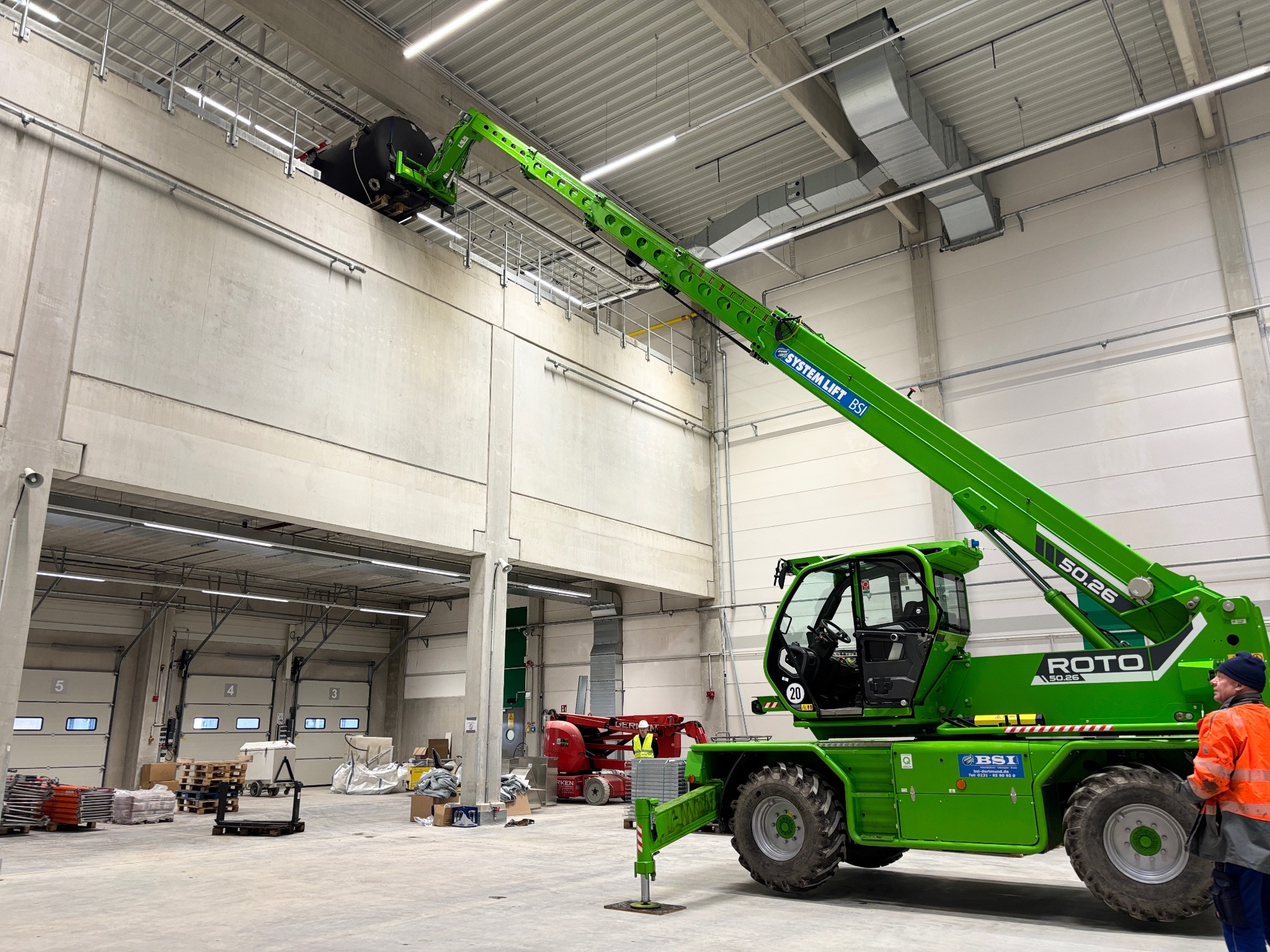 Montaż przemysłowej klimatyzacji w hali magazynowej za pomocą podnośnika teleskopowego. Widoczne elementy instalacji wentylacyjnej pod sufitem.