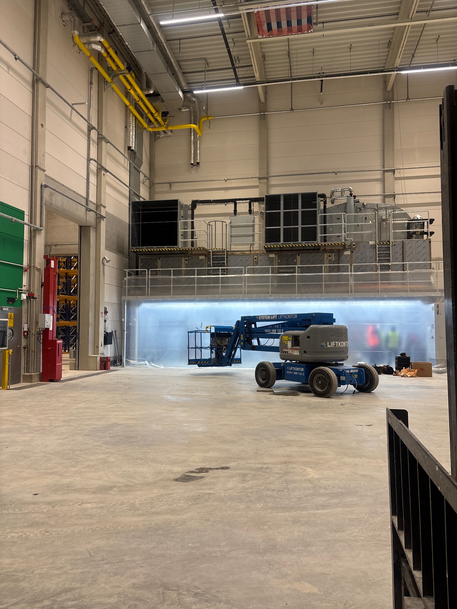 Montaż przemysłowej klimatyzacji w hali: podnośnik koszowy przed dużym agregatem, widoczne żółte rury wentylacyjne pod sufitem. Hala w trakcie prac instalacyjnych.