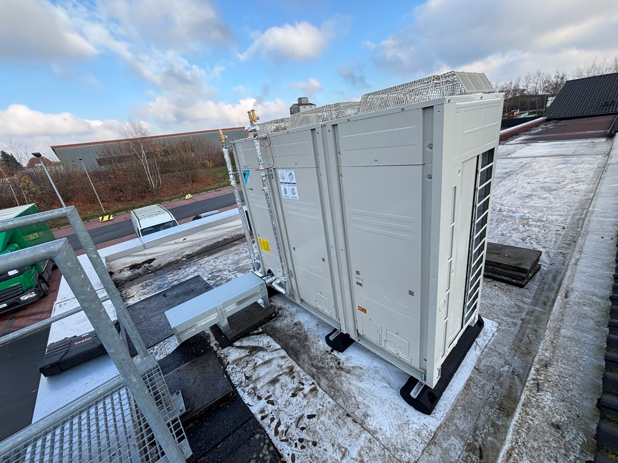 Biała, zewnętrzna jednostka klimatyzacji Daikin na dachu budynku, widoczna z perspektywy drabiny. Na dachu resztki śniegu, w tle budynek przemysłowy i błękitne niebo.