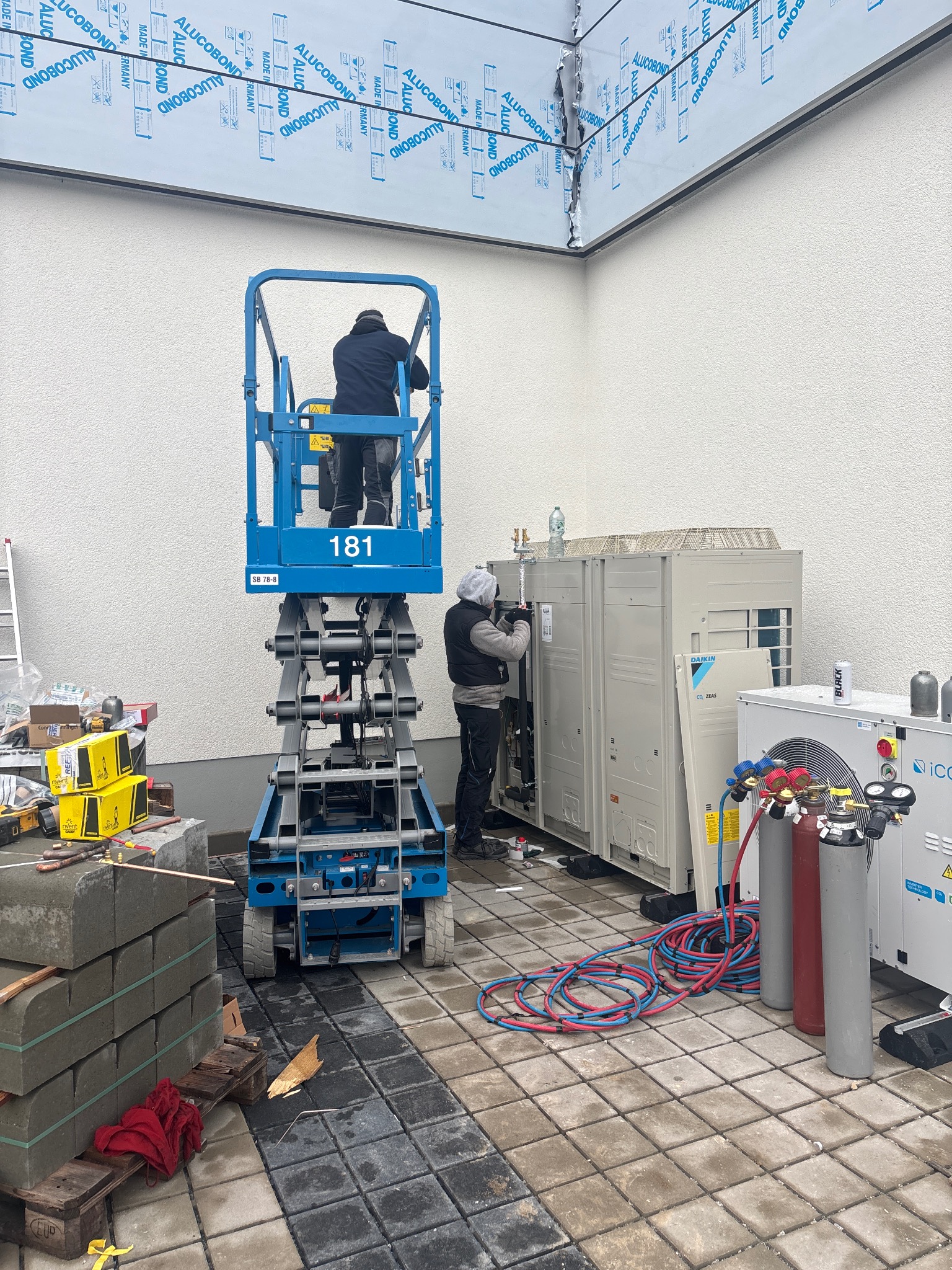 Montaż klimatyzacji: Dwóch monterów na podnośniku i przy jednostkach zewnętrznych Daikin CO2 ZEAS, butle z gazem, narzędzia na kostce brukowej. Praca w toku.