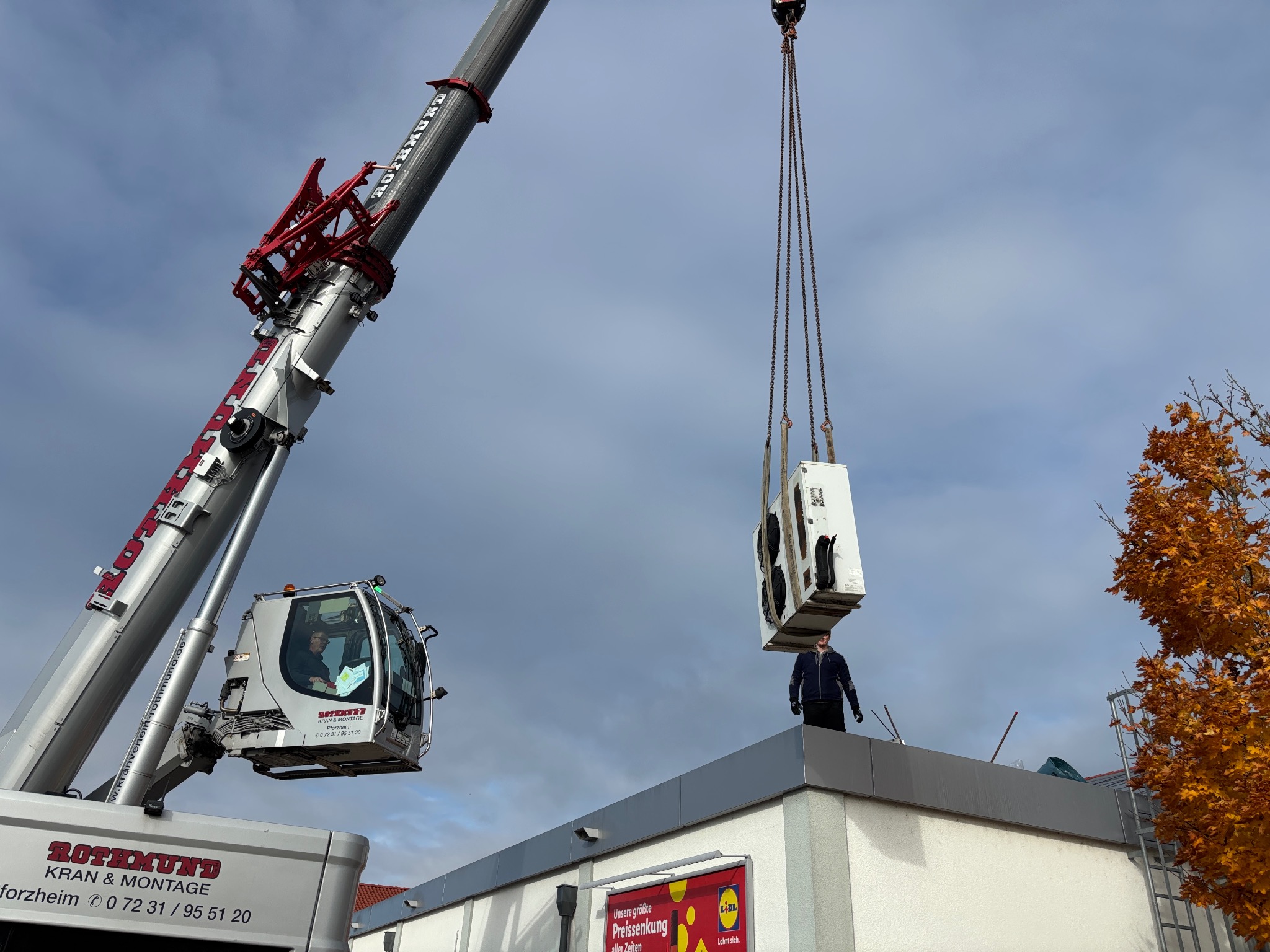 Montaż klimatyzacji na dachu budynku za pomocą dźwigu Rothmund. Widoczny operator w kabinie i pracownik na dachu. Elementy instalacji chłodniczej w zawieszeniu.
