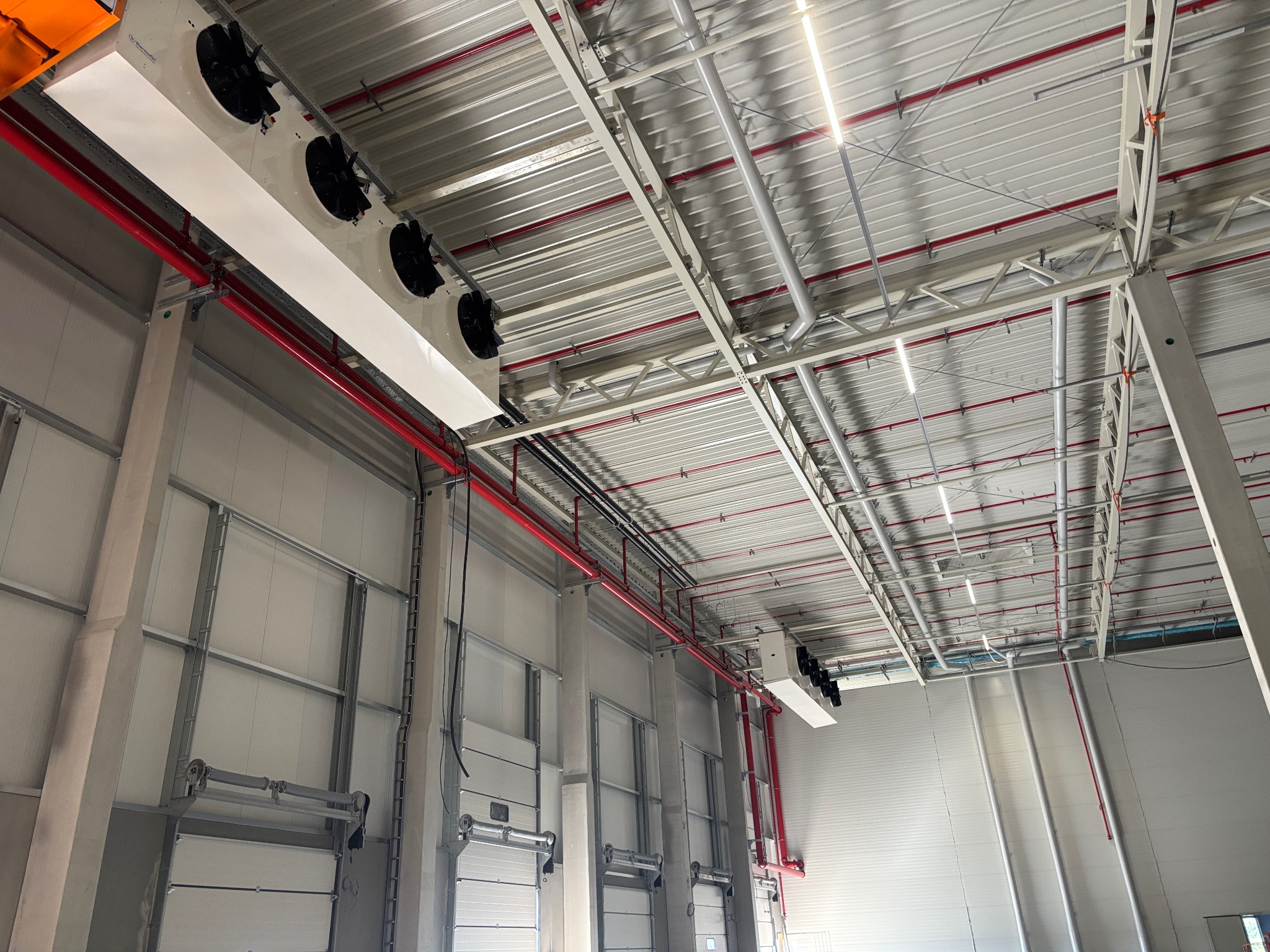 Zamontowana instalacja chłodnicza w hali magazynowej, widoczne agregaty chłodnicze pod sufitem, czerwone rury instalacji, metalowa konstrukcja dachu i bramy wjazdowe.