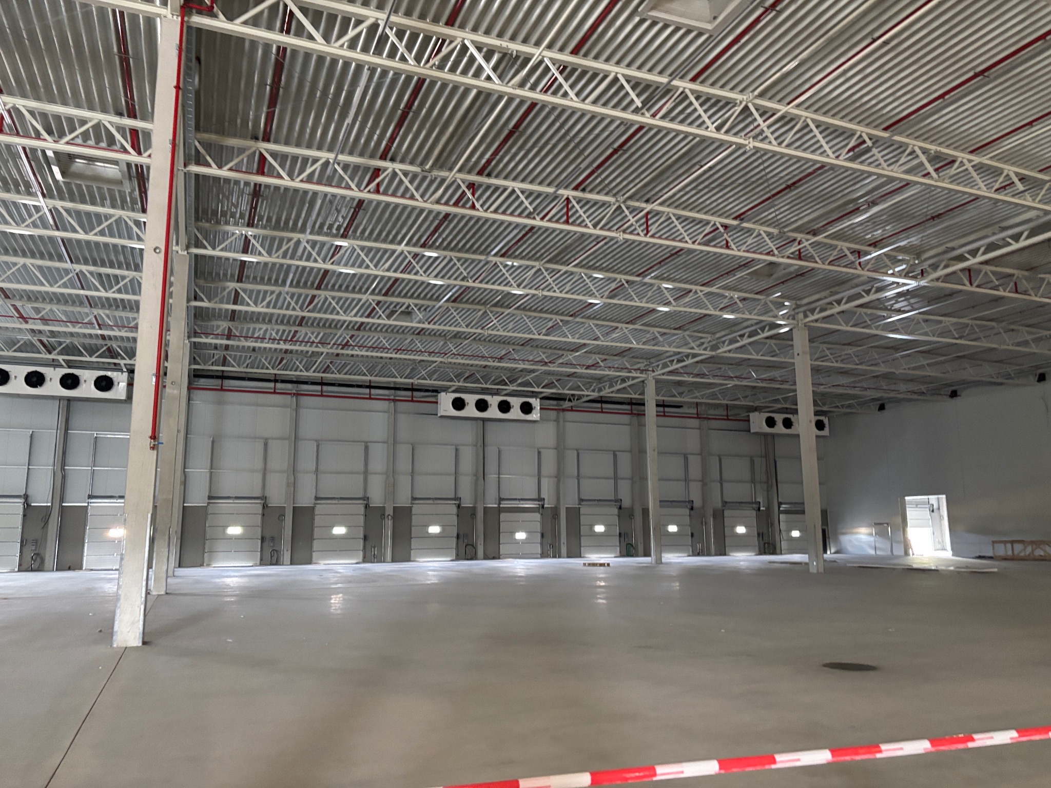 Wnętrze hali magazynowej z zamontowanymi agregatami chłodniczymi pod sufitem. Widoczne bramy wjazdowe i stalowa konstrukcja dachu z instalacjami.