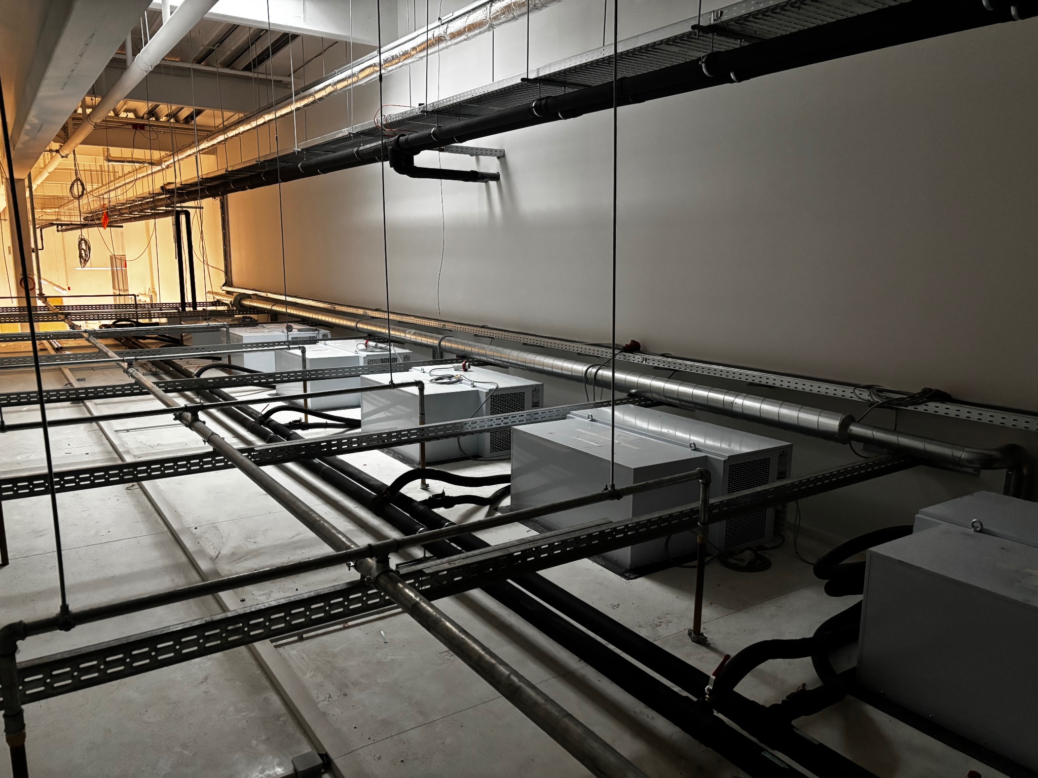 Instalacja chłodnicza w hali: szare agregaty, rury i kable na metalowych konstrukcjach pod sufitem. Widok z dołu, oświetlenie techniczne.