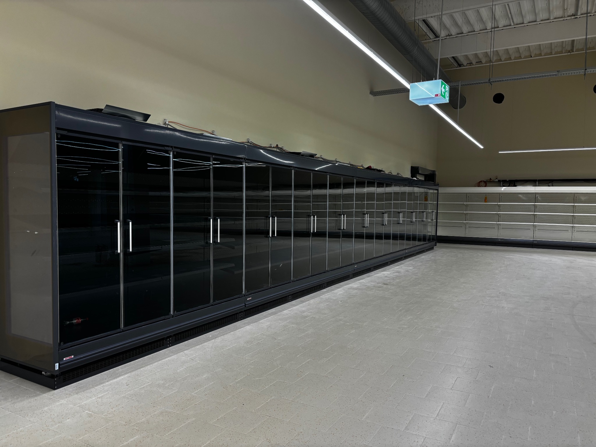Długa, pusta chłodnia z przeszklonymi drzwiami w sklepie, oświetlona liniowymi lampami LED. Widoczne puste półki w tle i betonowa podłoga.