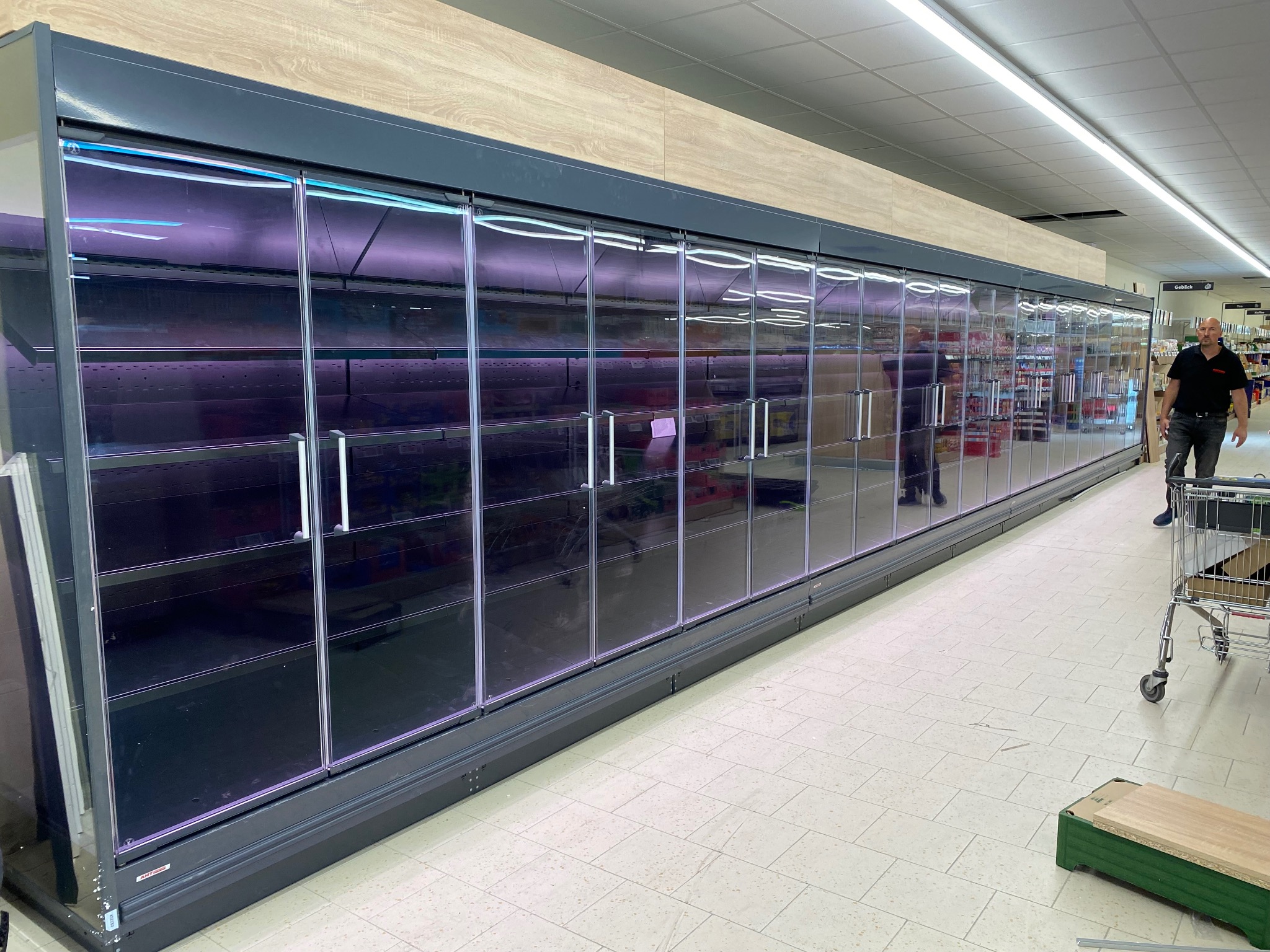 Długa, szara chłodnia z przeszklonymi drzwiami w supermarkecie, częściowo pusta, z widocznym fragmentem regałów i osobą w tle. Widoczny wózek sklepowy.