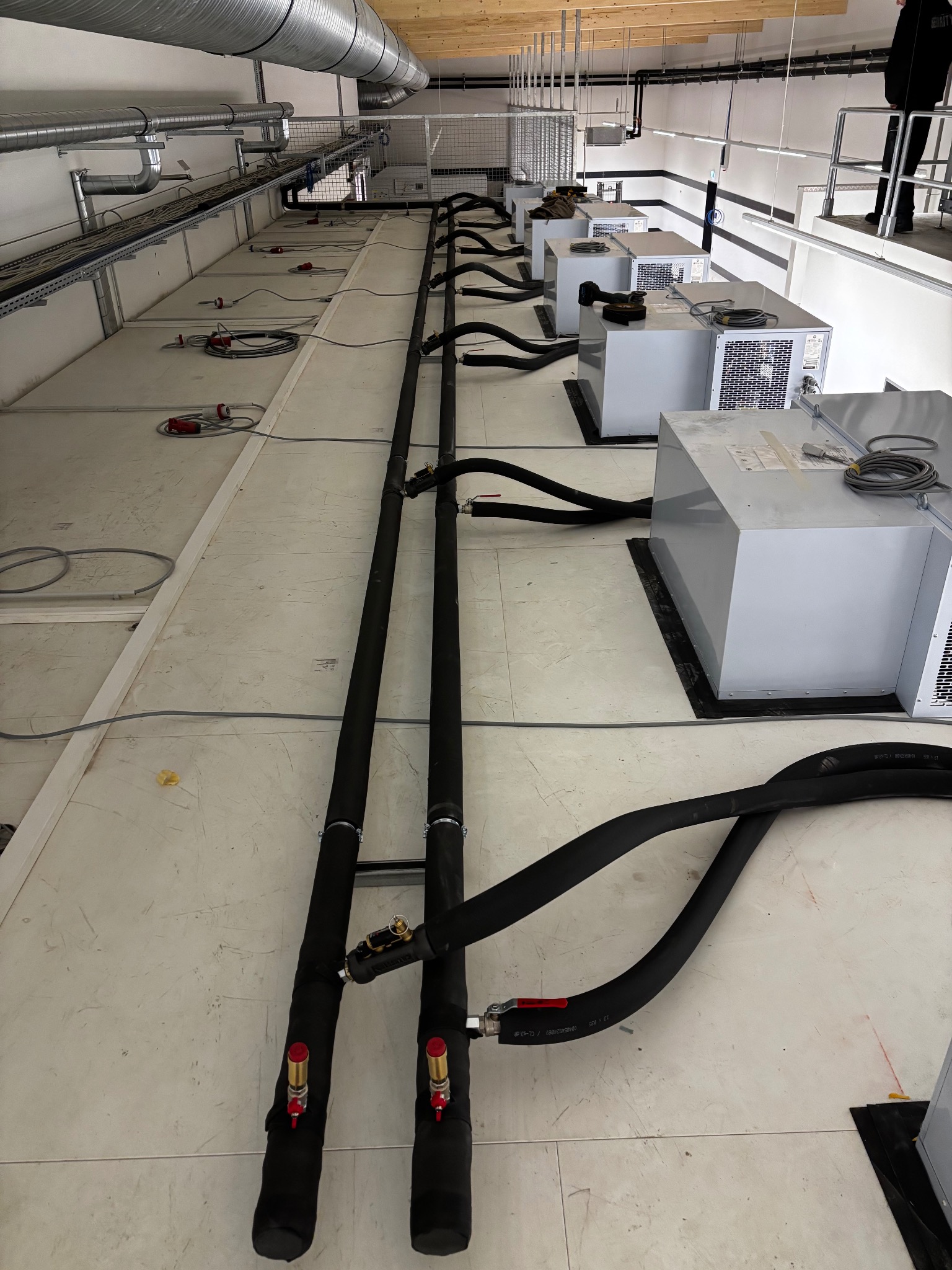 Instalacja chłodnicza w hali: rzędy agregatów z czarnymi rurami, kable w korytach pod sufitem, surowy stan wykończenia, widok z dołu pod kątem.