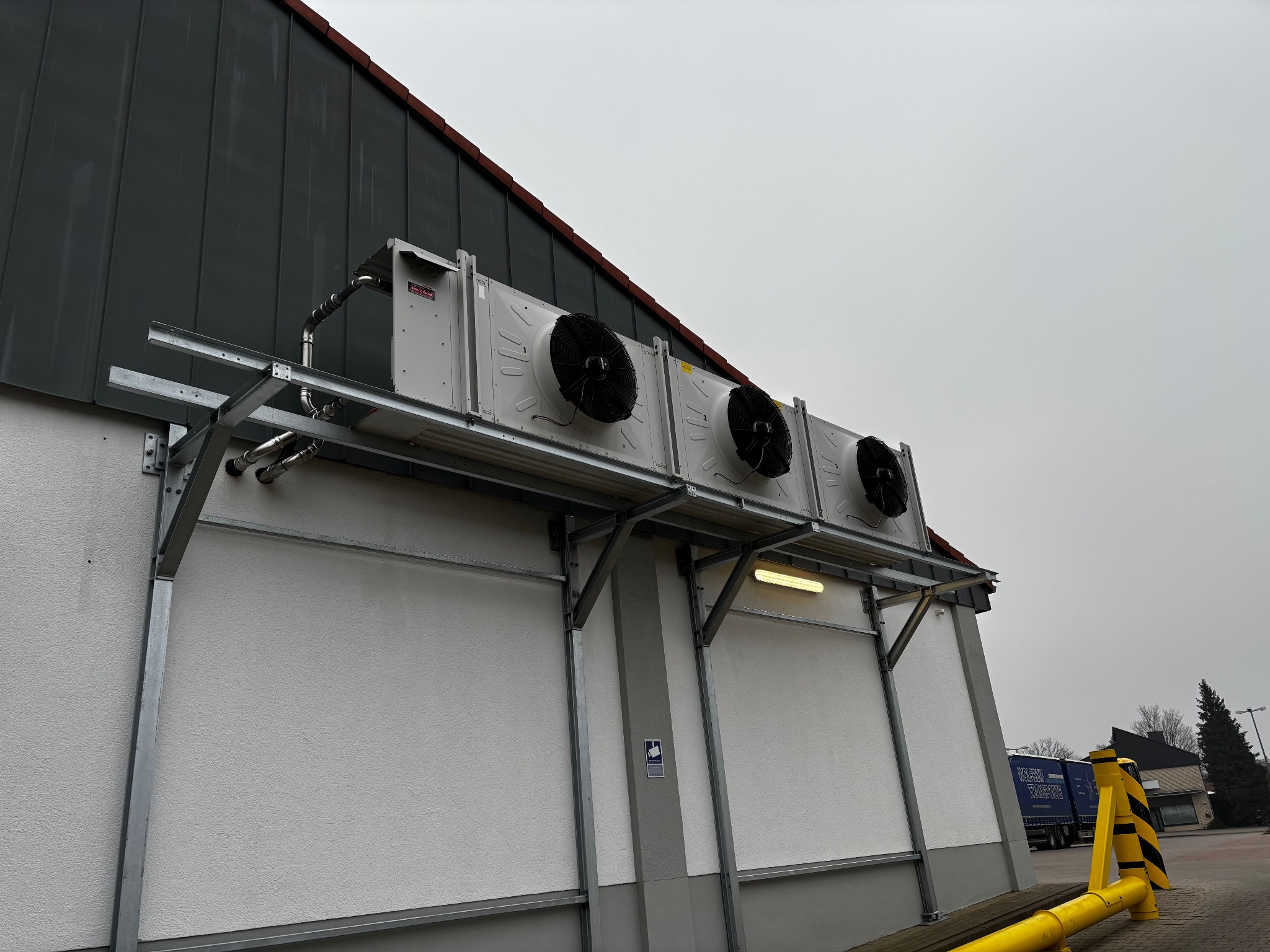 Zewnętrzne jednostki chłodnicze zamontowane na metalowej konstrukcji przy budynku z szarą elewacją. Widoczne rury i wentylatory. Pochmurne niebo w tle.